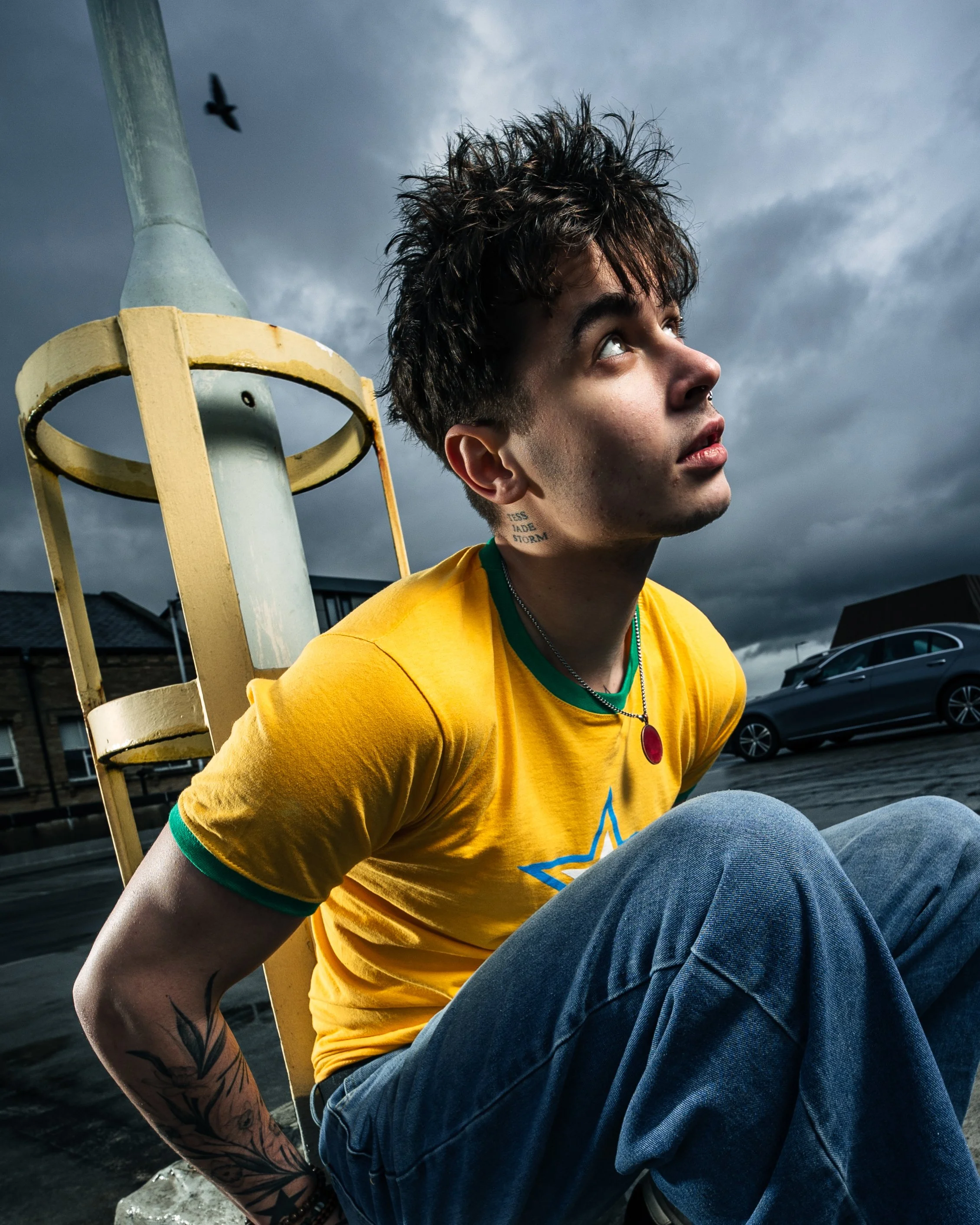 Young man with dark, spiky hair sitting on the ground, wearing a yellow T-shirt with a star design, blue jeans, and a necklace with a red pendant. He is looking upward under a cloudy sky, with a bird flying above and cars parked in the background.