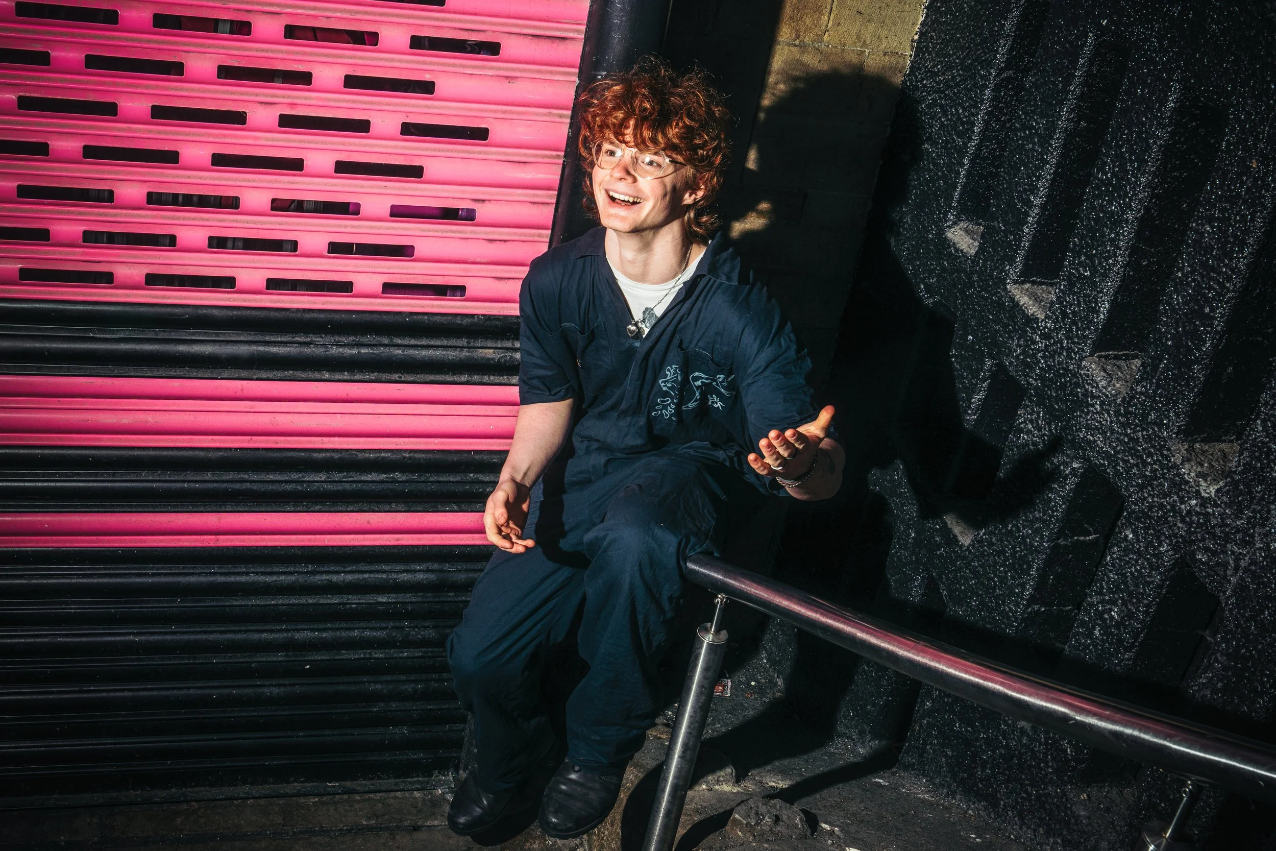 Young man with curly red hair wearing glasses and a dark blue jumpsuit, sitting on a ledge outside at night near a neon pink and black shutter door, gesturing with his right hand while smiling and looking up.