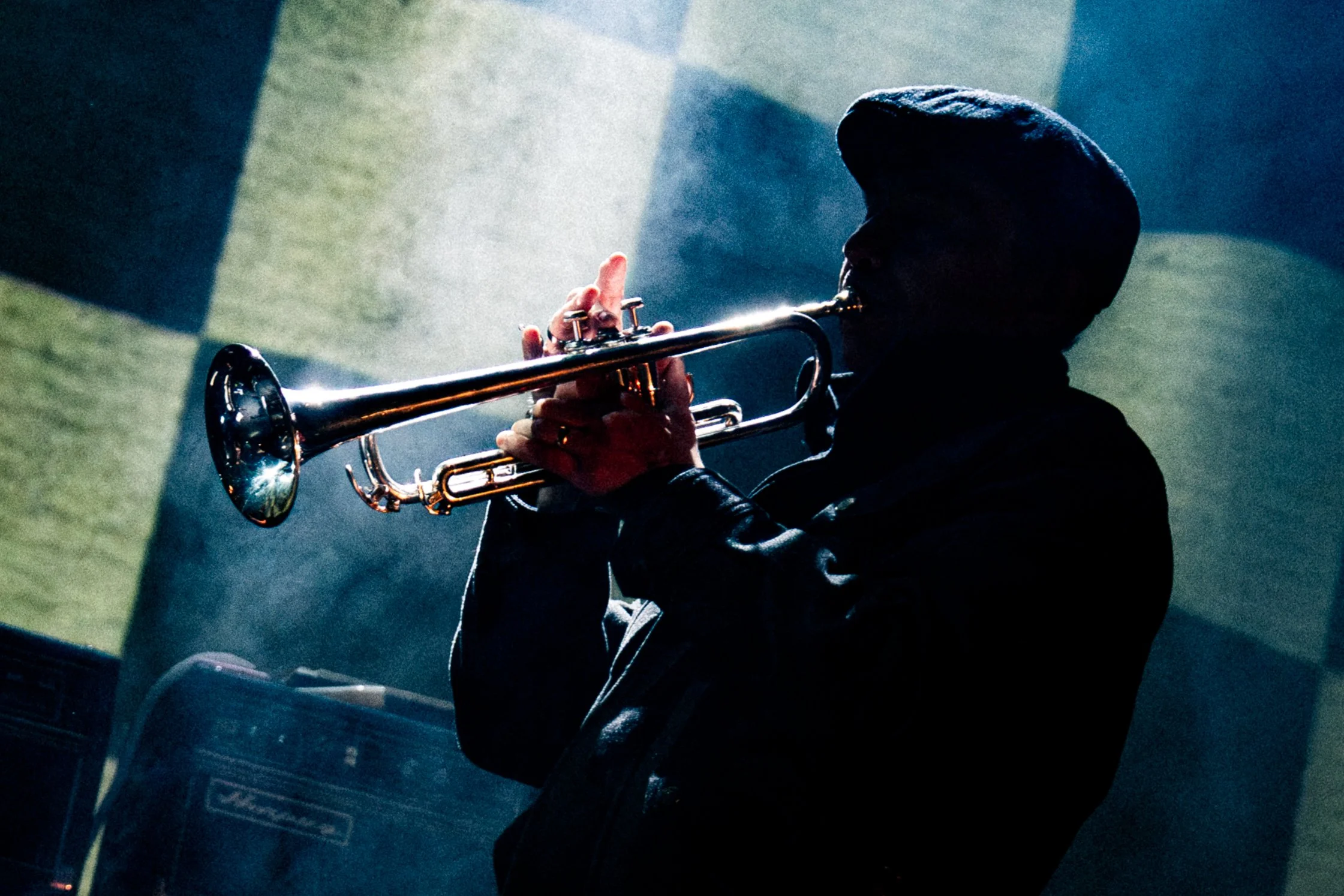 A person wearing a hooded jacket playing a trumpet in a dimly lit setting with colored banners and equipment in the background.