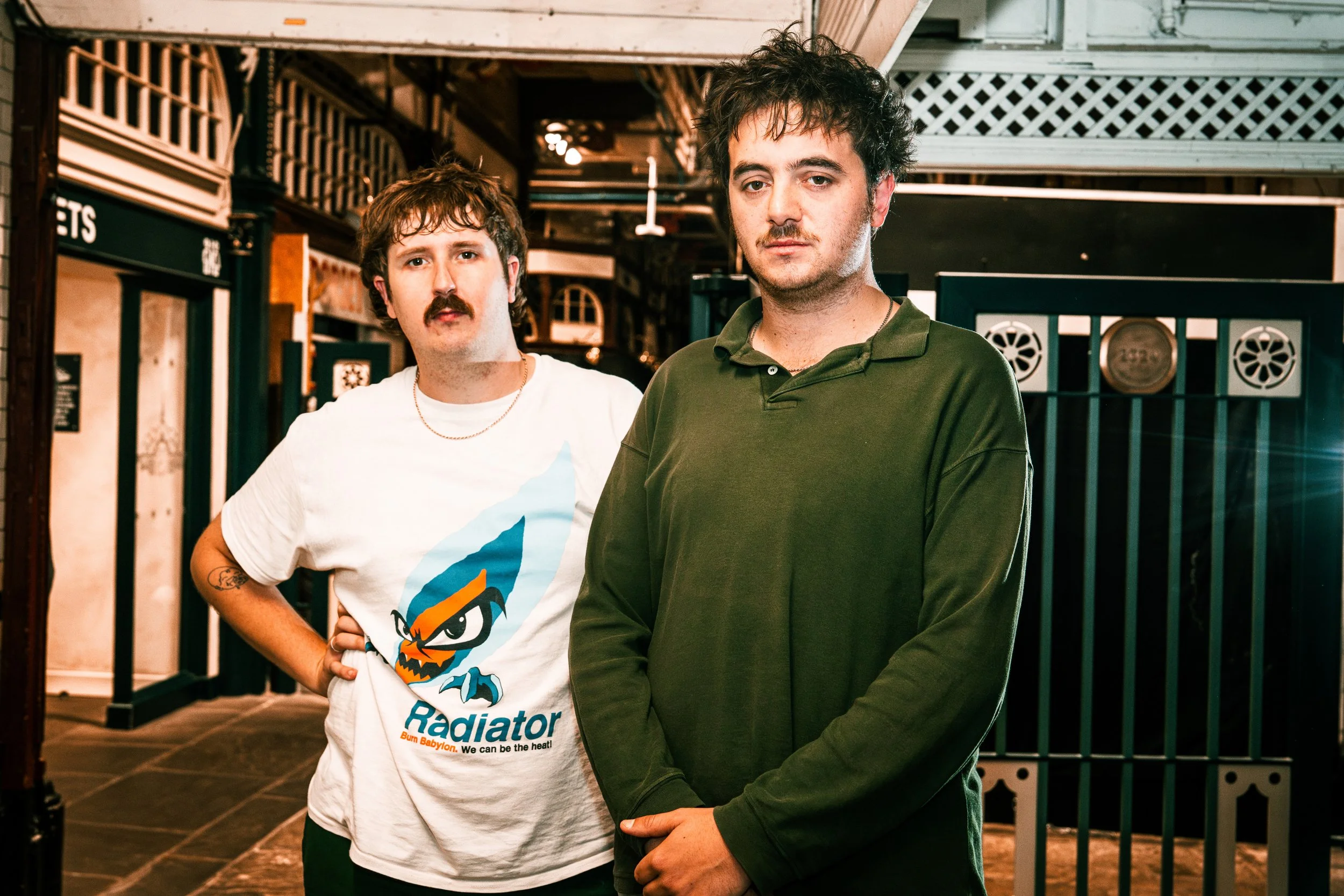 Two young men with curly hair and mustaches standing indoors against a dark background, one wearing a white T-shirt with a cartoon character and the other in a green polo shirt.
