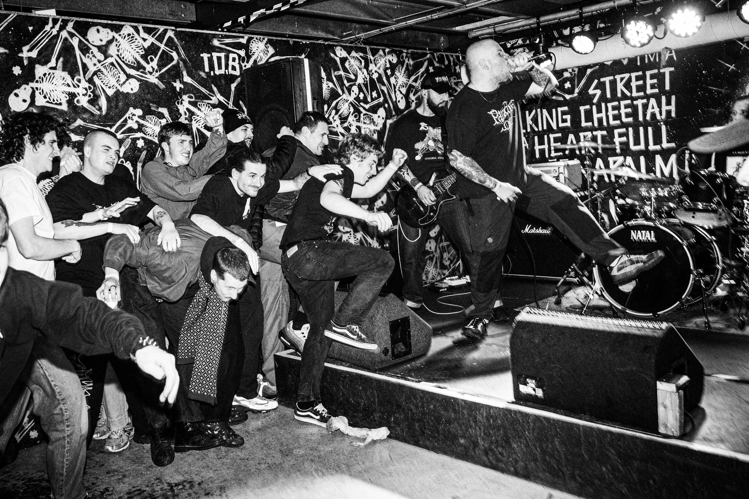 A black and white photo of a live music performance showing a lead singer with tattoos and a bald head in front of a crowd. The crowd is energetic, some members jumping and dancing. The stage has drums, a guitar, and a background sign with words like