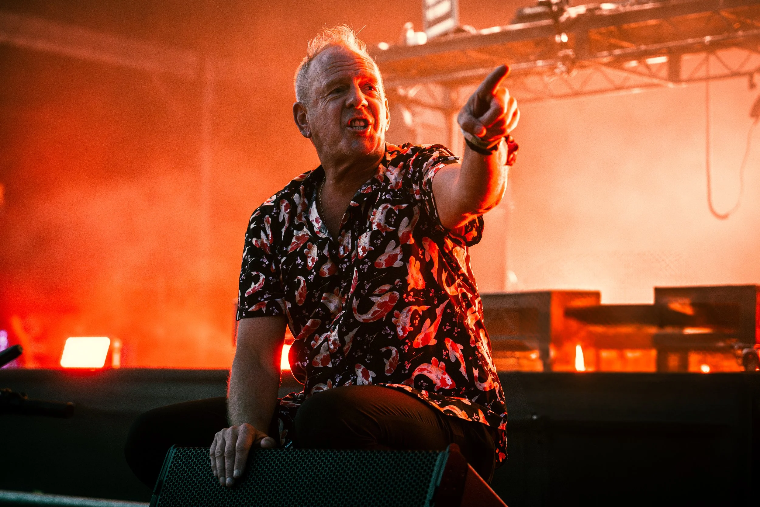 A man with blonde hair, wearing a colorful patterned shirt, sitting on a stage, pointing and speaking passionately with a bright, fiery red backdrop.