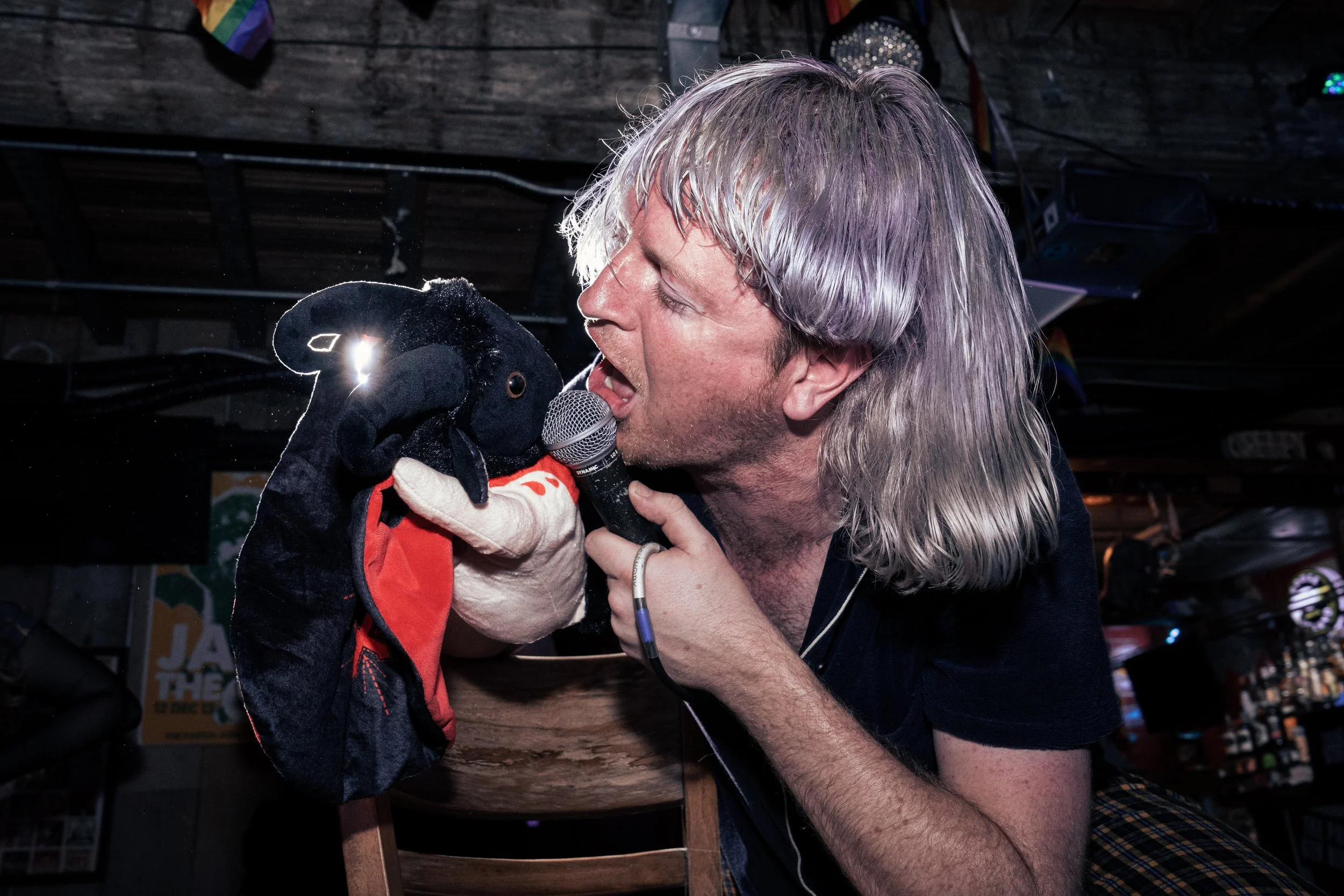 A man with long blonde hair sings into a microphone while holding a sock puppet of a black animal wearing a red scarf in a dimly lit bar or pub.