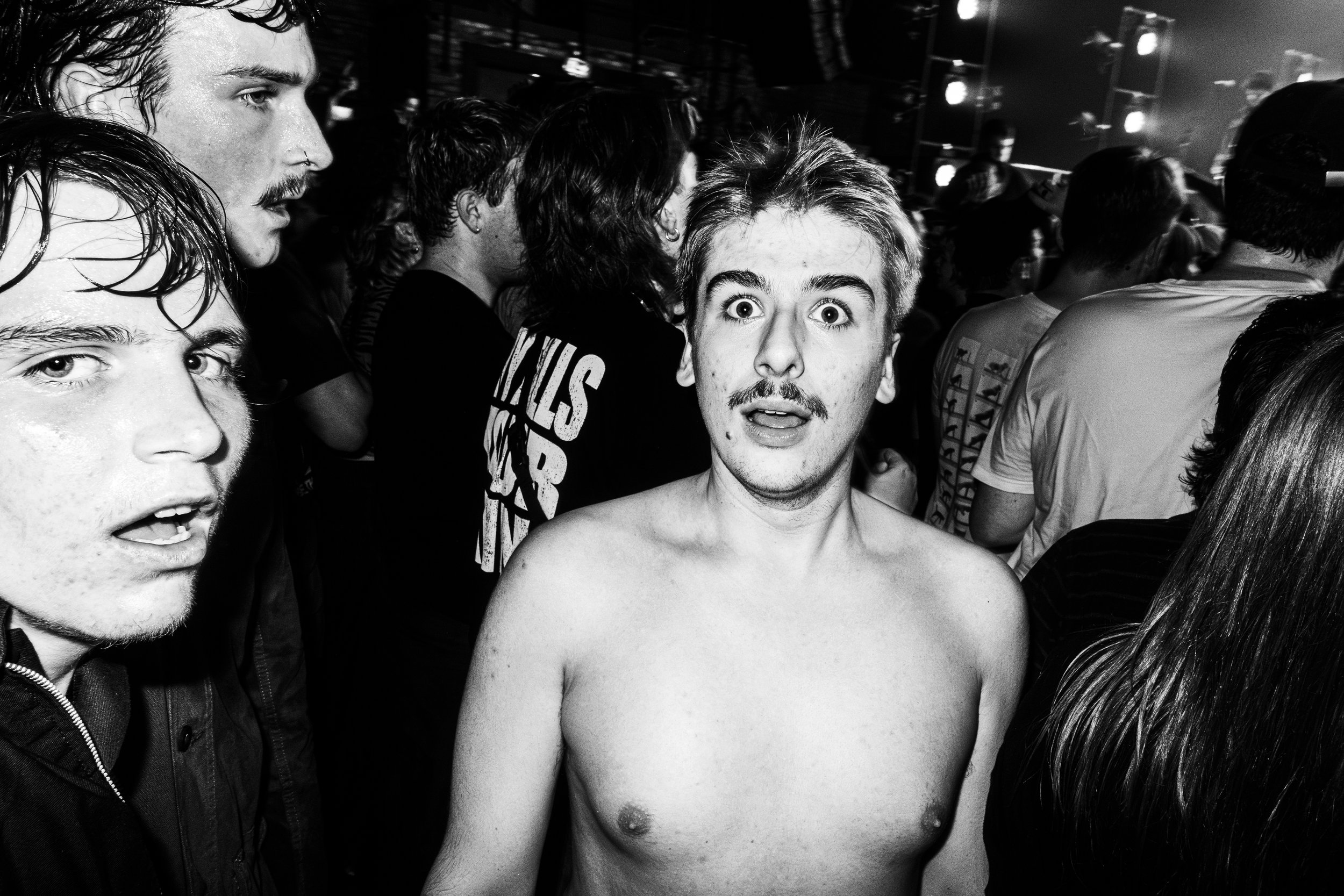 A young man with a mustache and short hair is shirtless and standing among a crowd at a concert or club, with an surprised or shocked expression on his face. The crowd around him includes other young people, some with wet hair, and the background sho