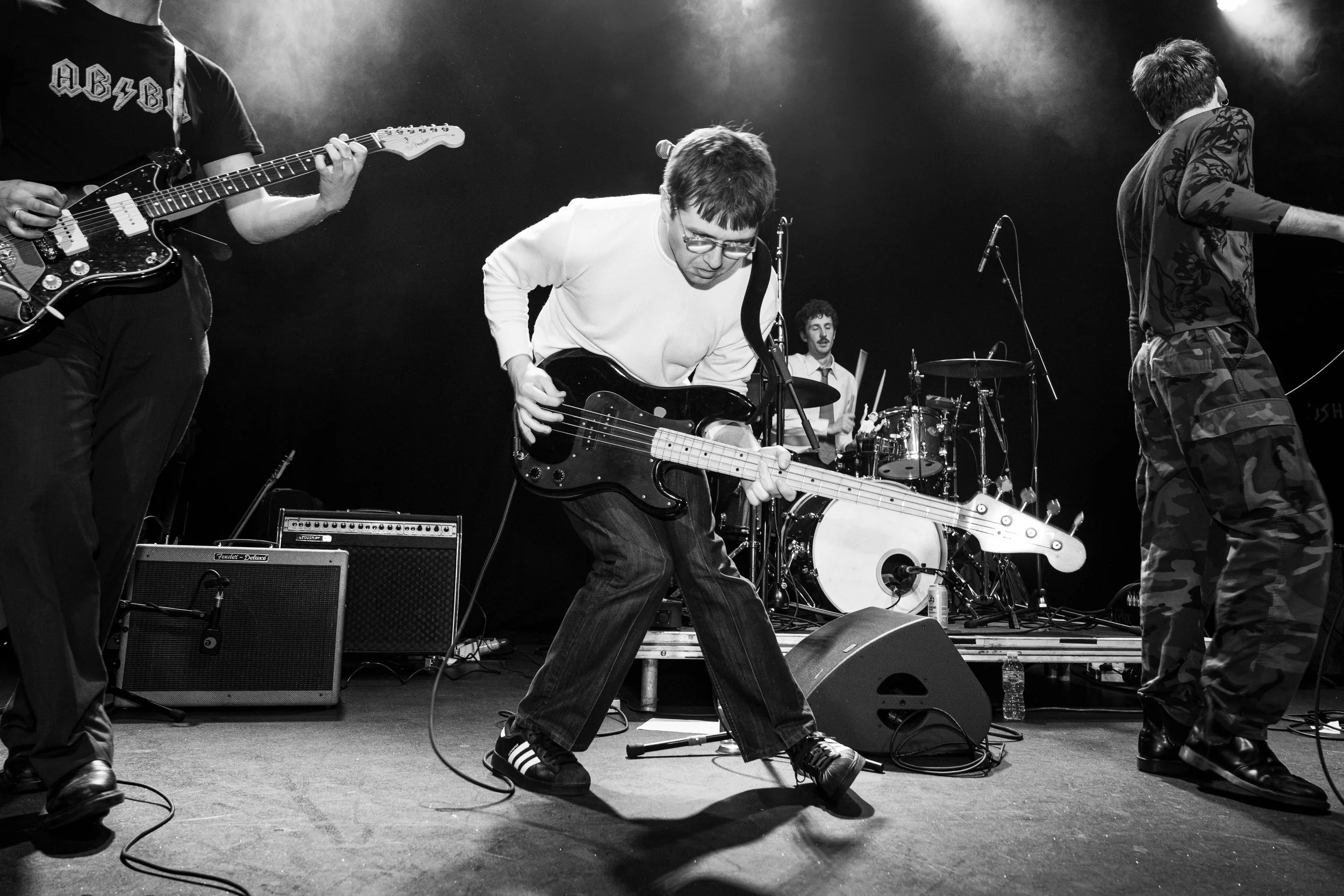 A black-and-white photo of a band performing live on stage. One musician in the center is playing an electric bass guitar with a focused expression, wearing glasses and a white sweater. To the left, a guitarist is playing with part of his body visibl