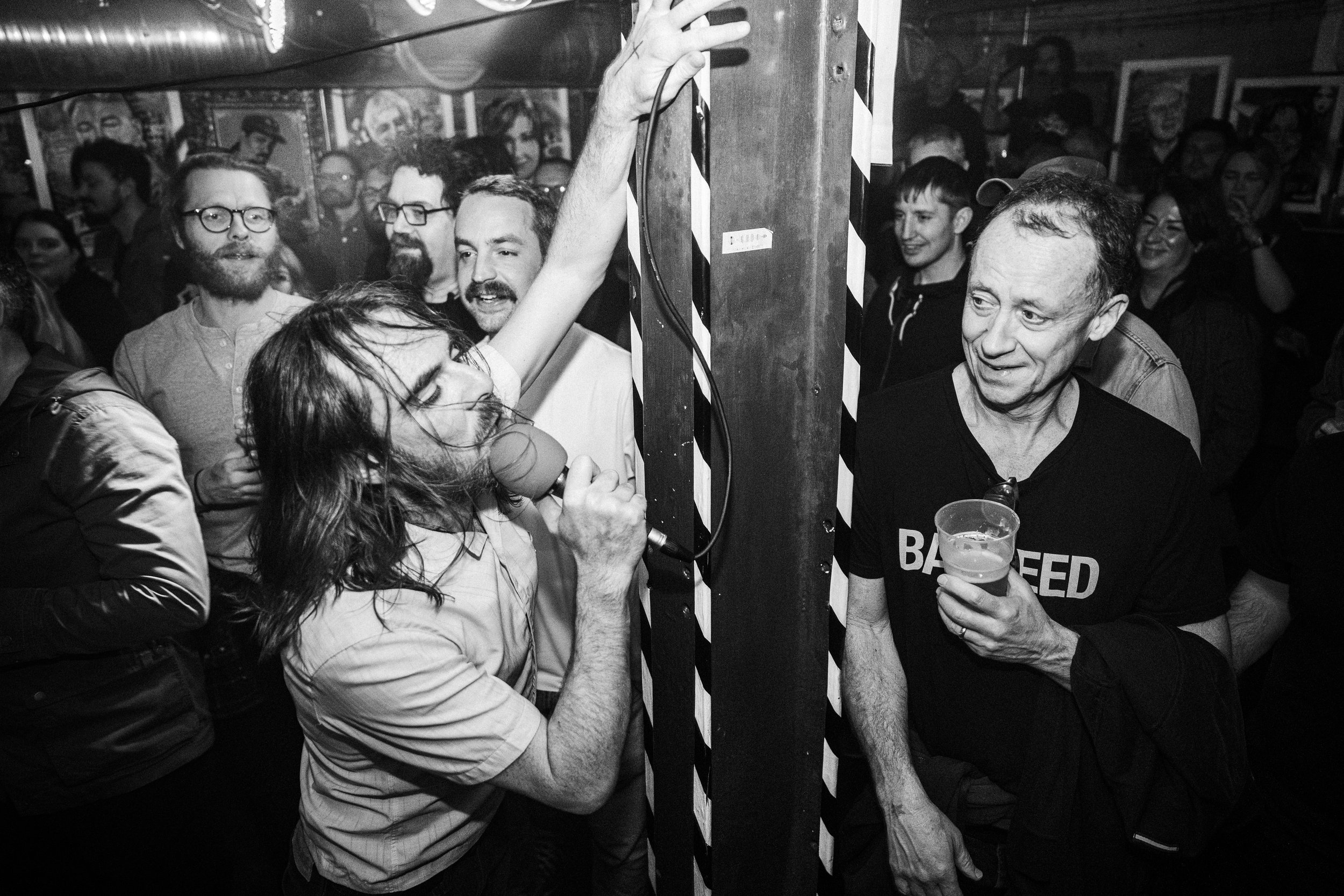 Group of people at a lively event, with a woman singing into a microphone on the left and a man holding a drink on the right. Some are smiling and others are watching.