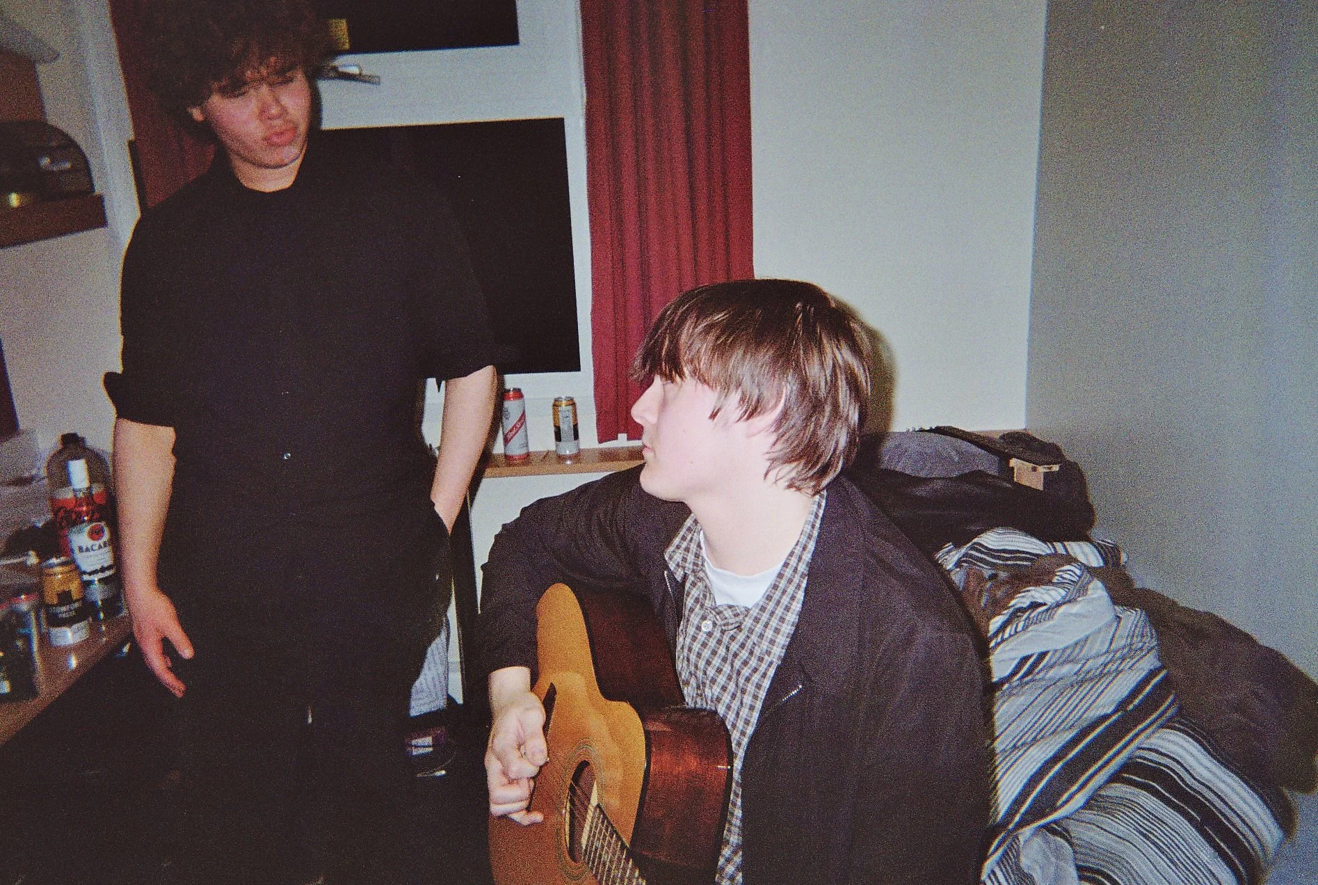 Two young men in a room, one sitting playing an acoustic guitar with a plaid shirt and black jacket, the other standing wearing a black shirt, with a TV, cans, and a bed with striped bedding in the background.