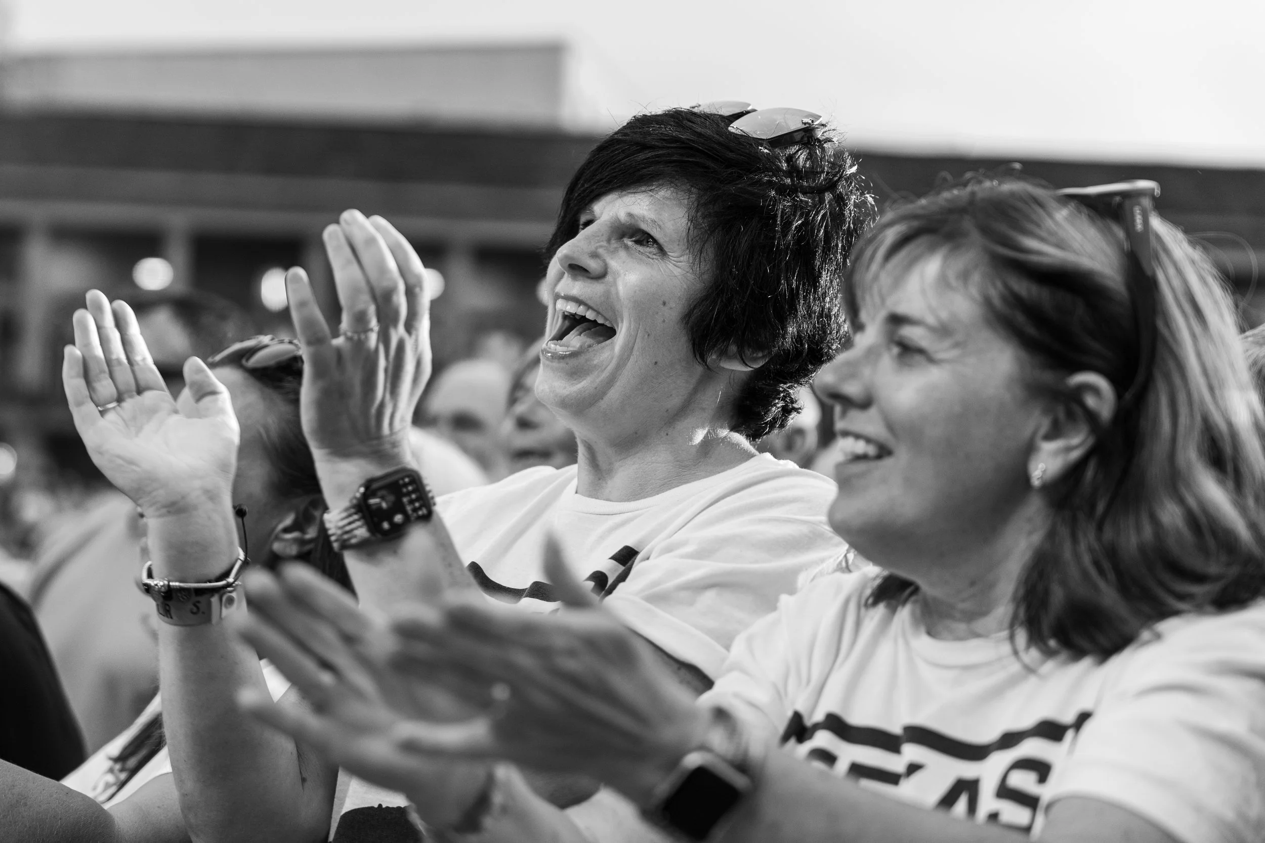 Two women, smiling and clapping, at an indoor event, wearing casual white shirts with text, one with short dark hair and glasses on her head, the other with shoulder-length hair and earrings, captured in a lively moment.