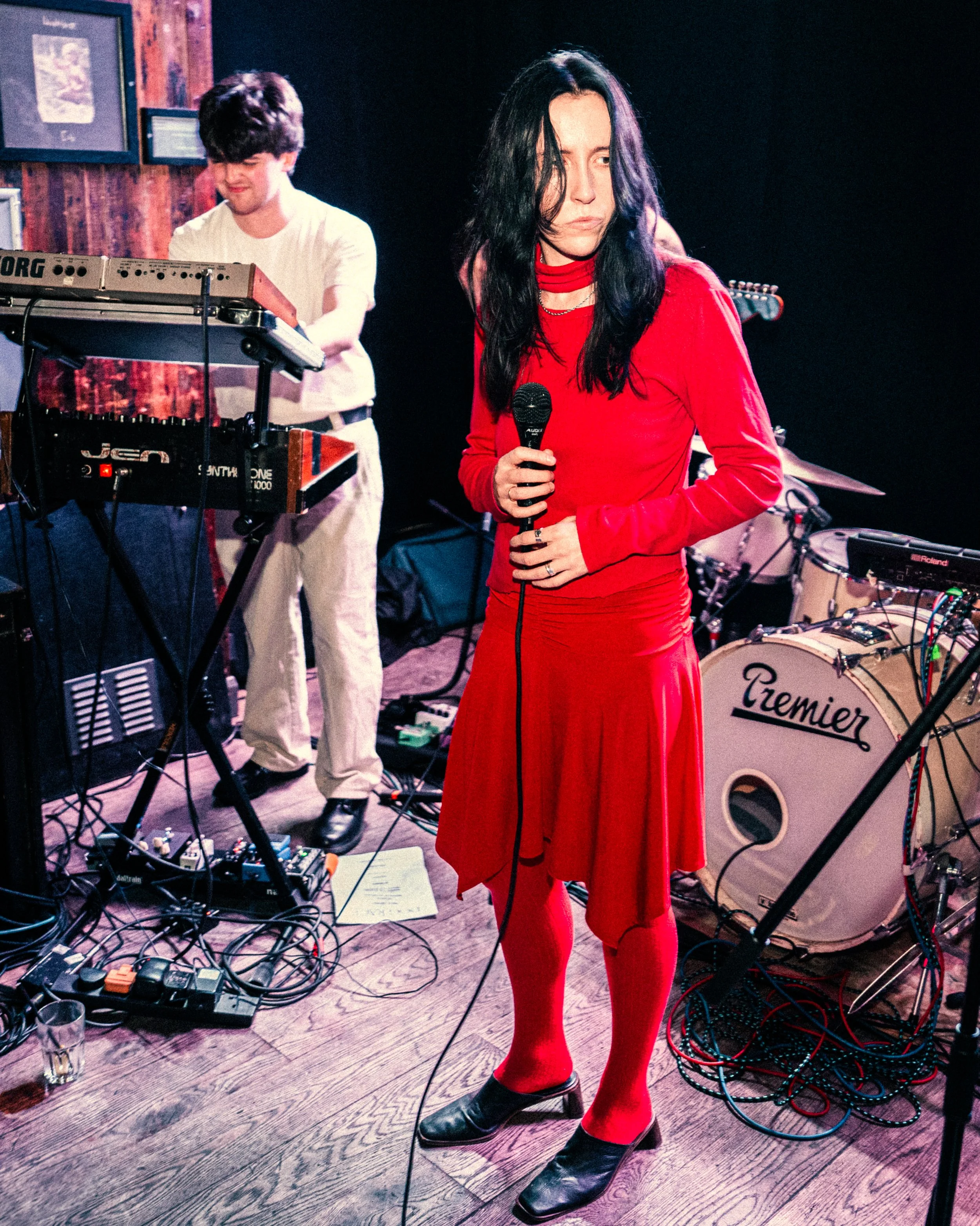 A woman with long dark hair in a red dress and red tights holds a microphone on stage, with a keyboardist and drum set in the background.