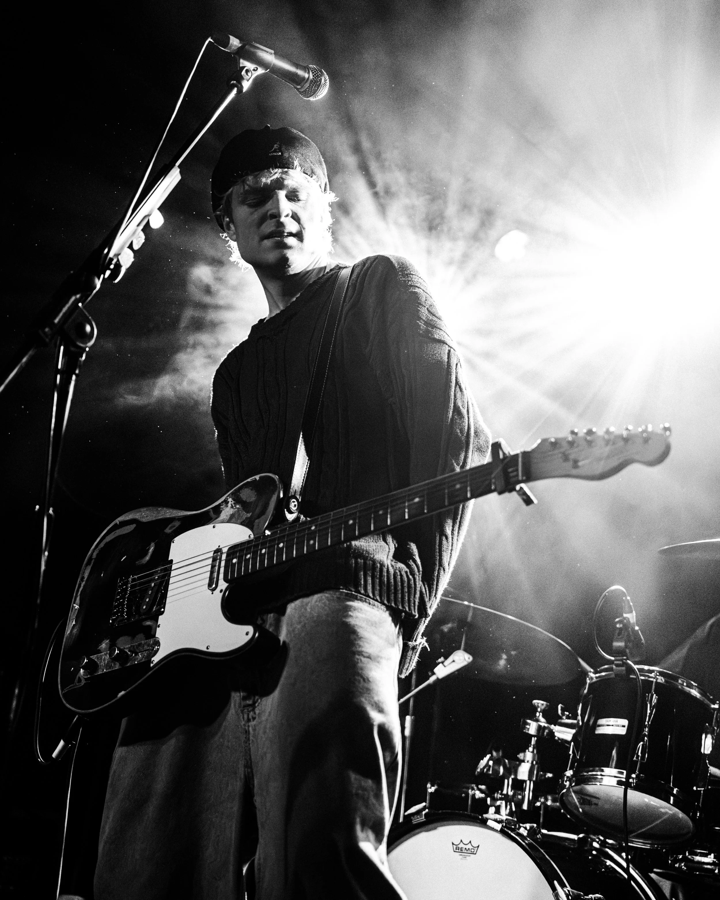 Black and white photo of a musician playing guitar on stage with bright light behind him, microphone in foreground, and drum set in background.