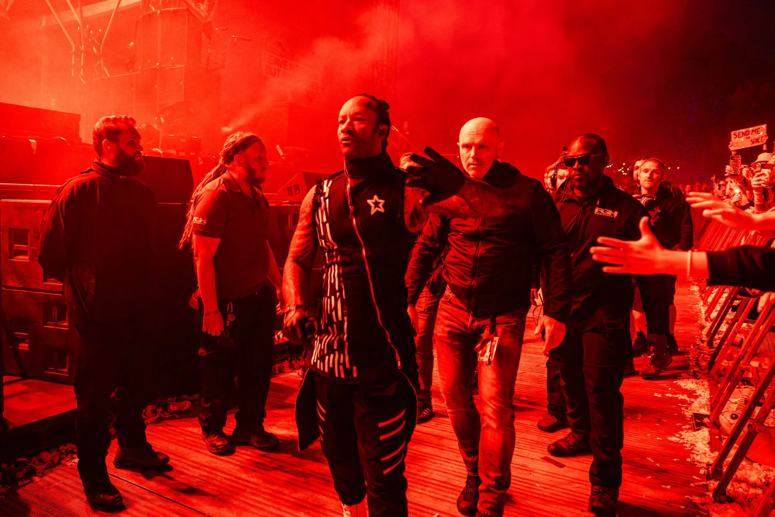A group of security personnel and staff walking through a concert or event venue with red lighting and smoke effects, with audience members visible on the right side.
