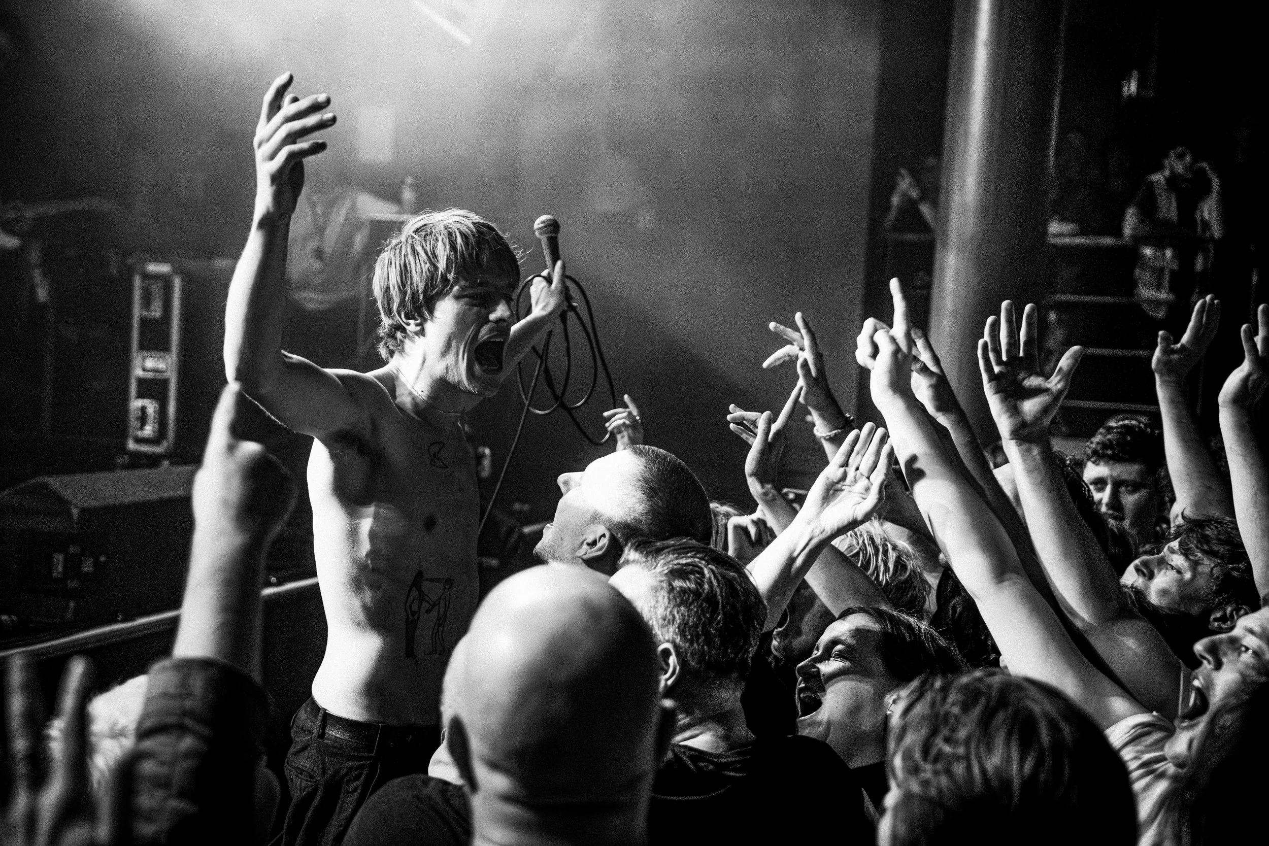 Black and white photo of a shirtless male lead singer with blonde hair performing passionately on stage with a microphone, surrounded by an audience reaching towards him with their hands raised.