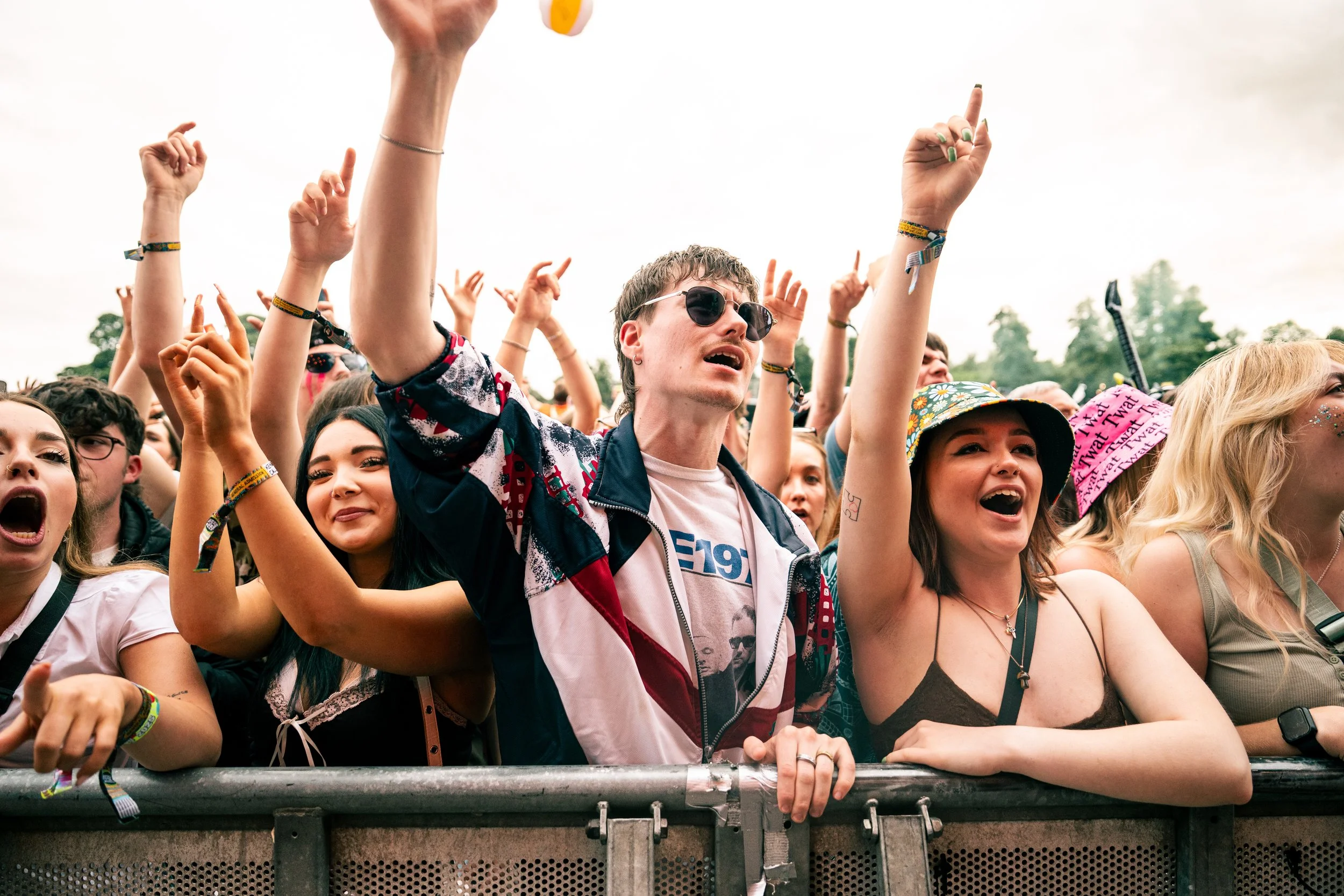 Crowd of people at a concert or music festival, smiling, cheering, and raising their hands near the stage.