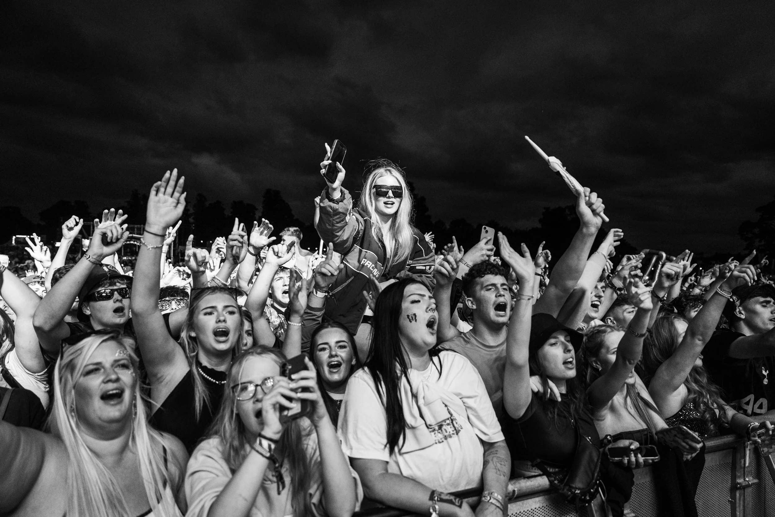 Crowd of diverse people at a concert or outdoor event, cheering and raising hands, with dark cloudy sky in background.