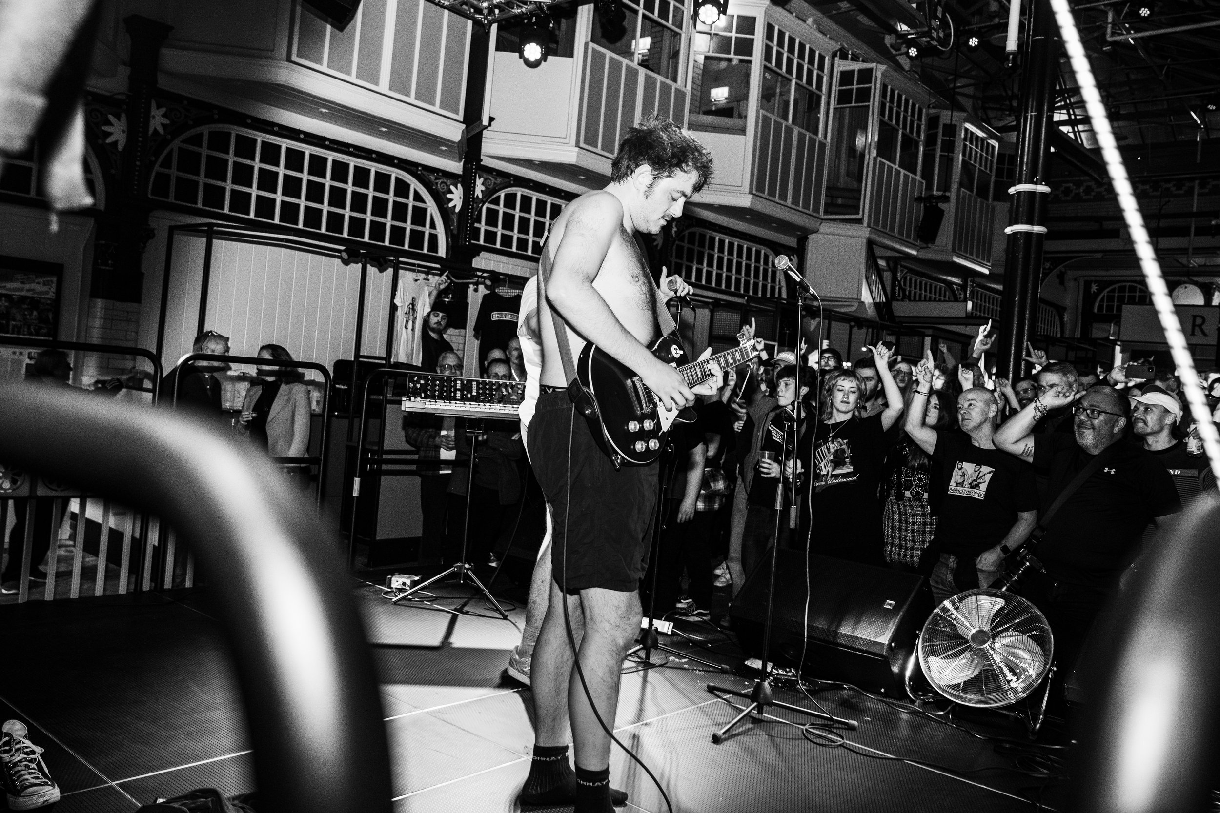 A musician with tousled hair, shirtless, wearing shorts, playing an electric guitar on stage. The crowd is dancing and enjoying the performance in an indoor venue with balconies and decorative architecture.