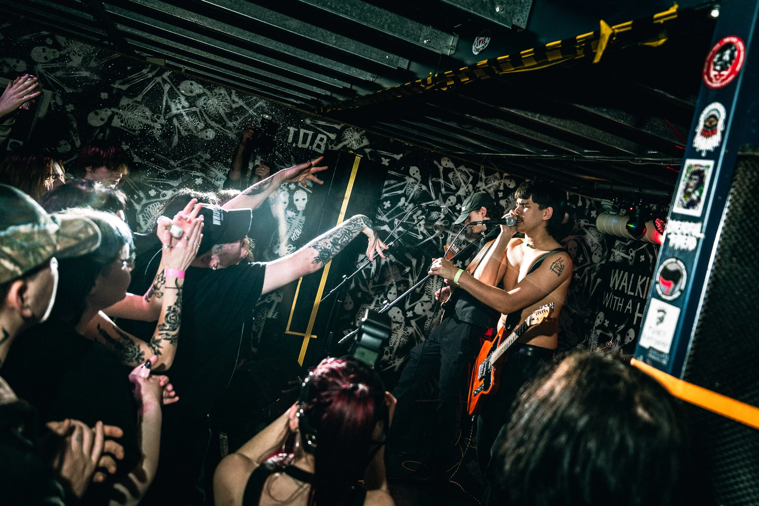 A live music performance at a small concert venue with a male singer playing an electric guitar and singing into a microphone, surrounded by an audience reaching toward him. The venue has black walls with white skeleton and butterfly decals.