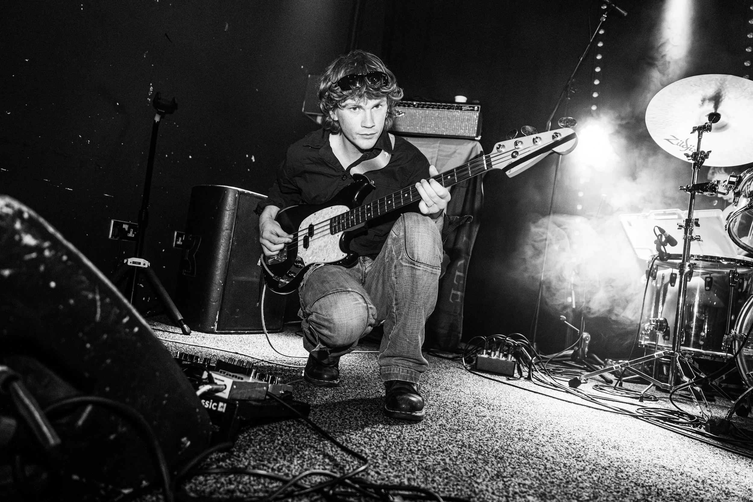 A musician crouching on stage playing an electric bass guitar, surrounded by musical equipment, with dramatic lighting from behind, in a black-and-white photo.