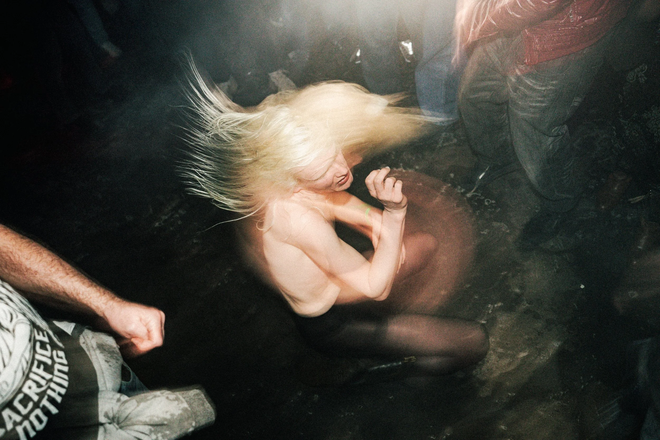 A person with long blonde hair dancing passionately on a dark, possibly muddy or dusty floor in a crowded space, with others standing around.