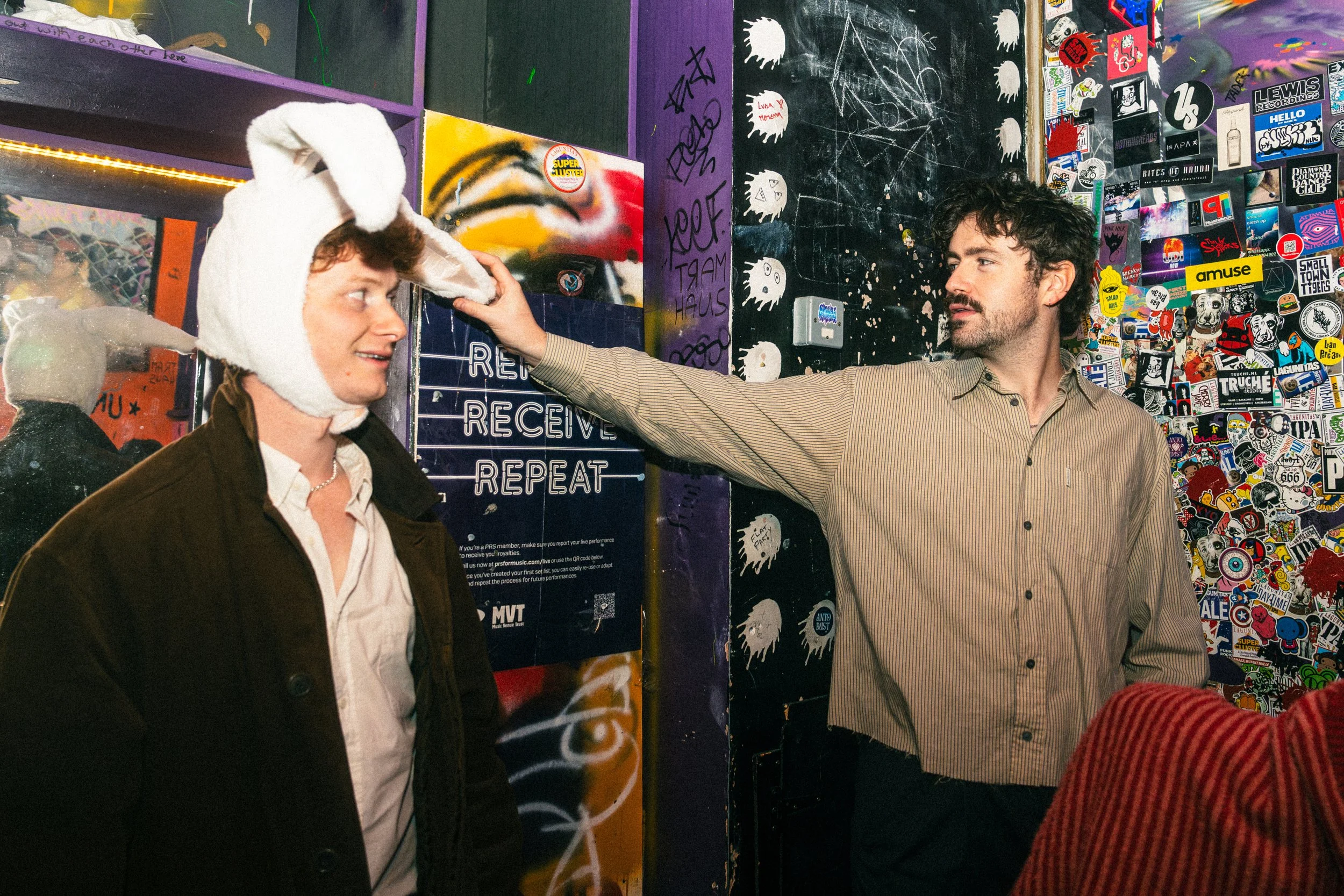 Two men indoors, one wearing a white animal costume hat and a brown jacket, the other in a striped shirt with facial hair, reaching out to touch the costume hat. The background is filled with colorful graffiti, stickers, and posters.