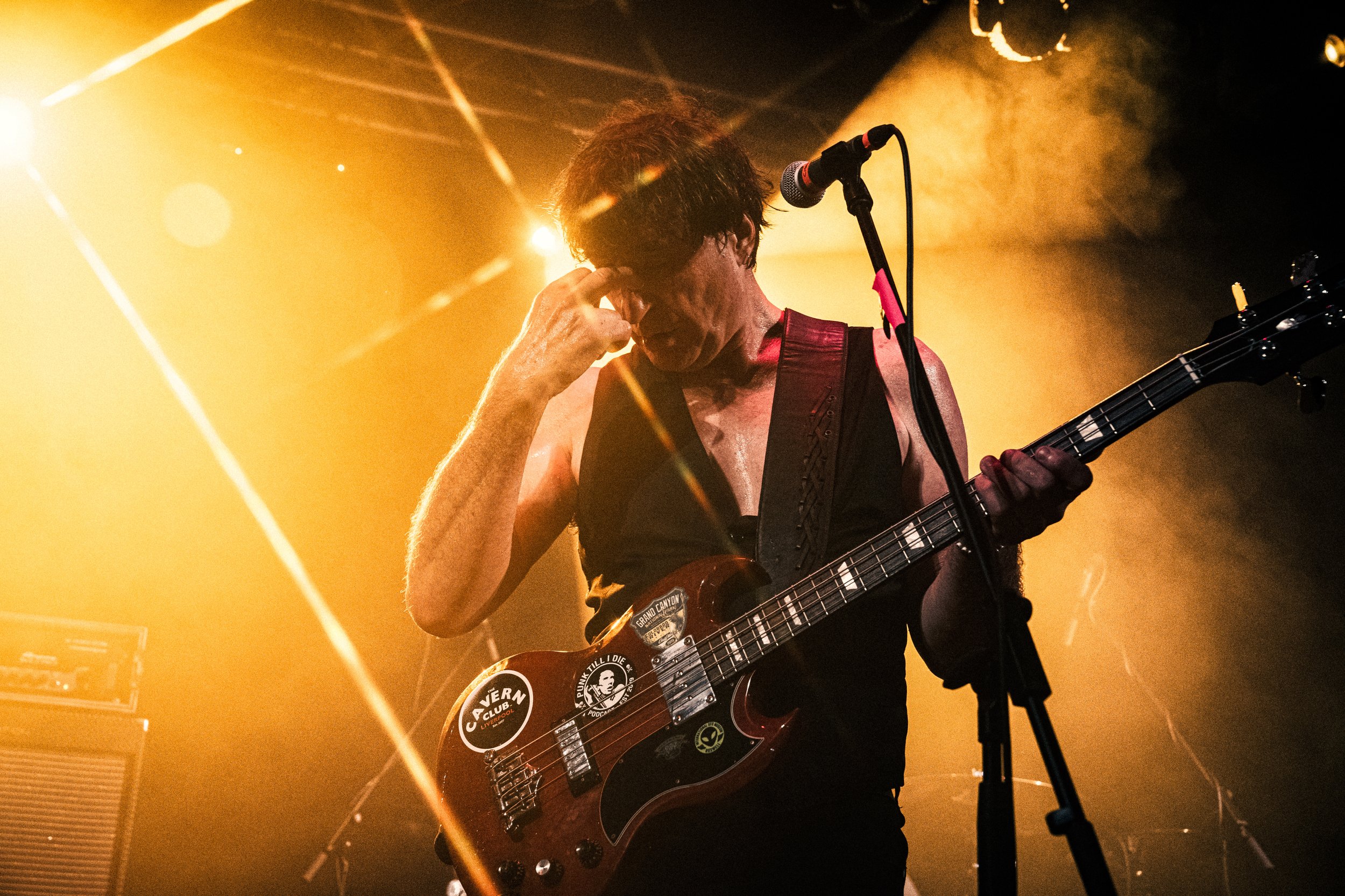 A male guitarist performing on stage with a guitar covered in stickers, under warm yellow stage lights, with a microphone in front of him.