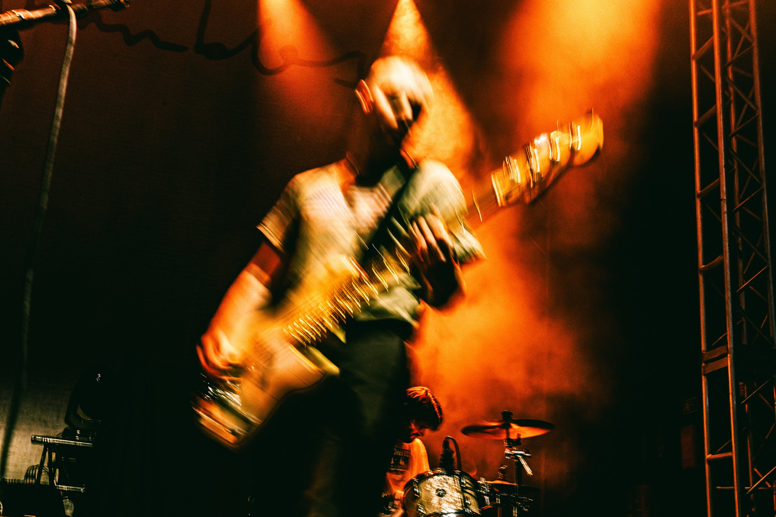 Musician playing electric guitar on stage with orange lighting, with a drummer in the background.