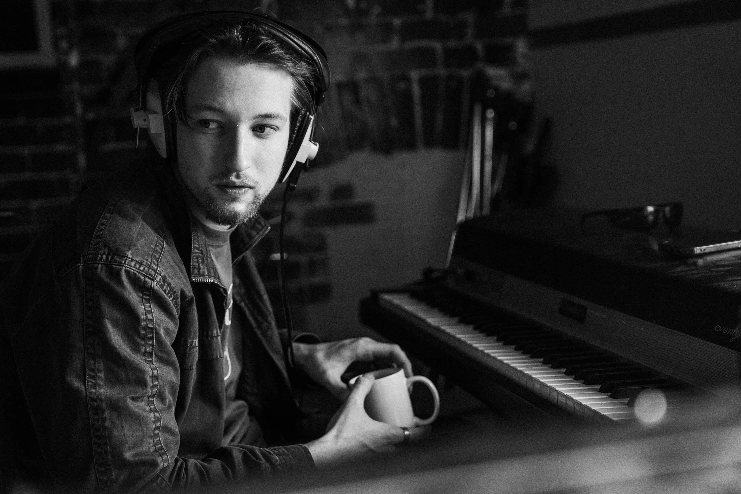 A young man wearing headphones sitting at a piano, holding a mug, looking away from camera in a dimly lit room with brick walls.