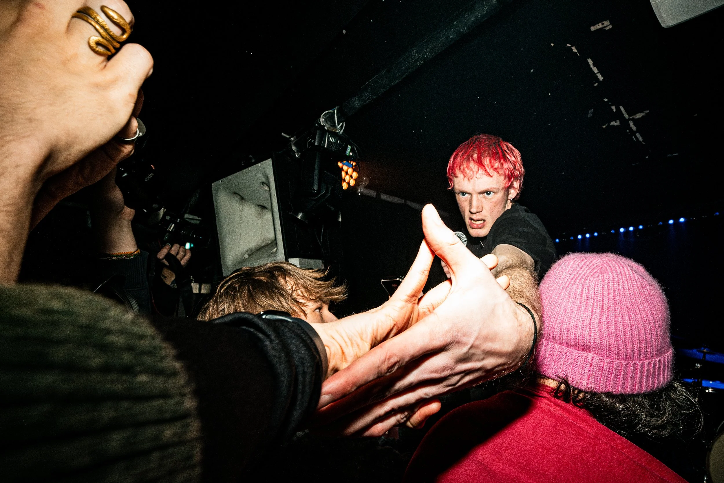 A performer with pink hair at a concert or event, interacting with the crowd and reaching out to someone with a finger in the foreground.