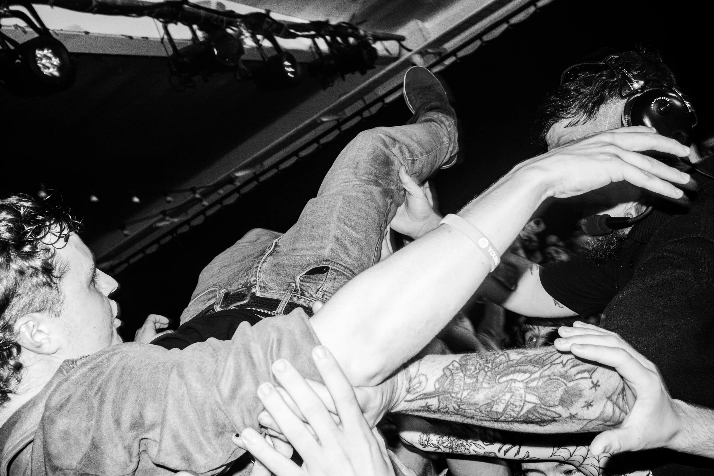 A chaotic scene at a concert or event shows people reaching and grabbing at a man in jeans and a black t-shirt, who is wearing headphones and leaning over a microphone, with the audience's hands touching him from all sides.