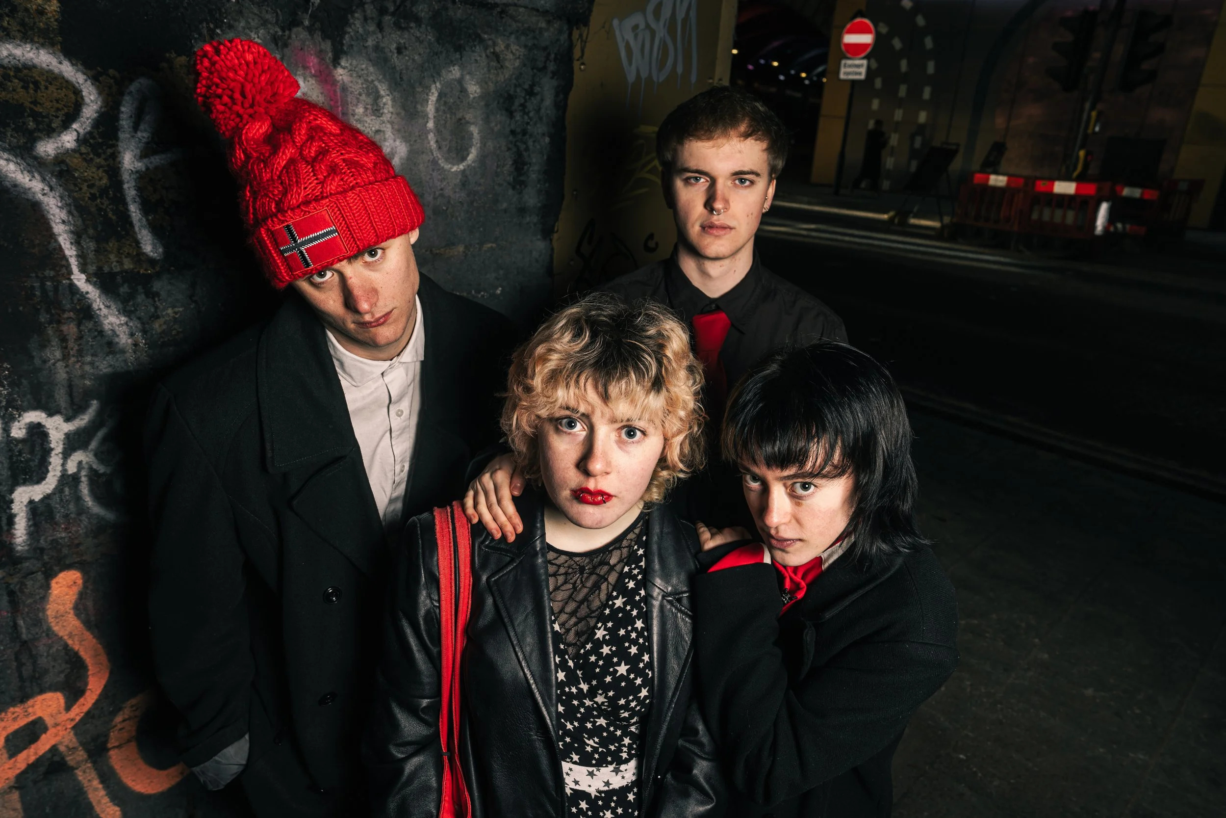 Four young adults standing close together in an urban setting at night, against a graffiti-covered wall, with a dark street and no entry sign in the background, dressed in dark fashion with red accents.