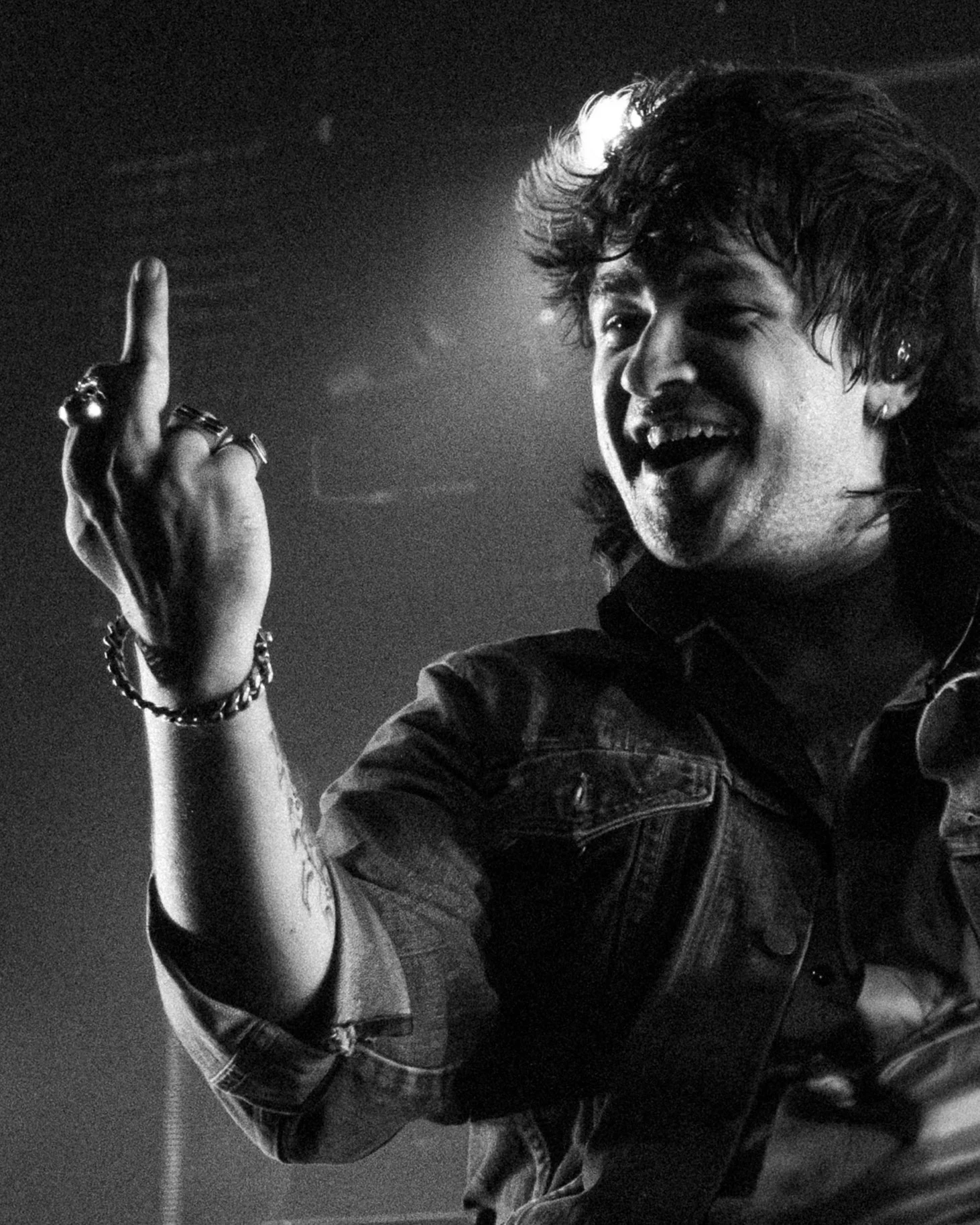 Black and white photo of a young man smiling with his mouth open, wearing a denim jacket, jewelry, and making a gesture with his raised hand.