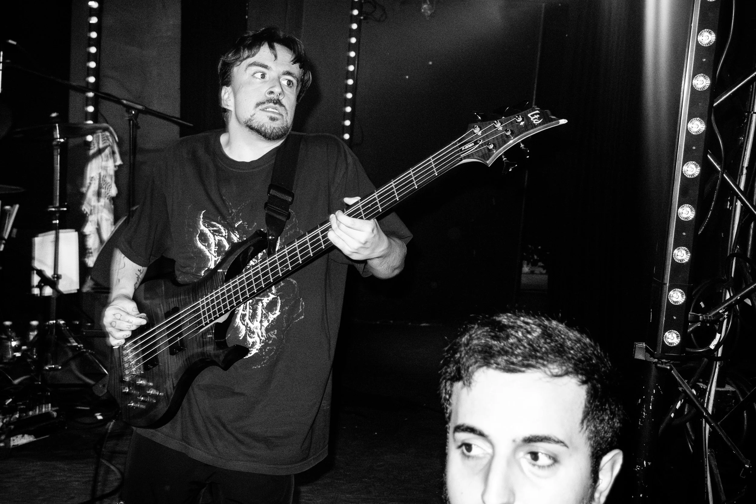 A man with a beard and mustache playing an electric bass guitar on stage, appearing focused with wide eyes, wearing a dark T-shirt with a skull graphic, while another person's head is in the foreground.