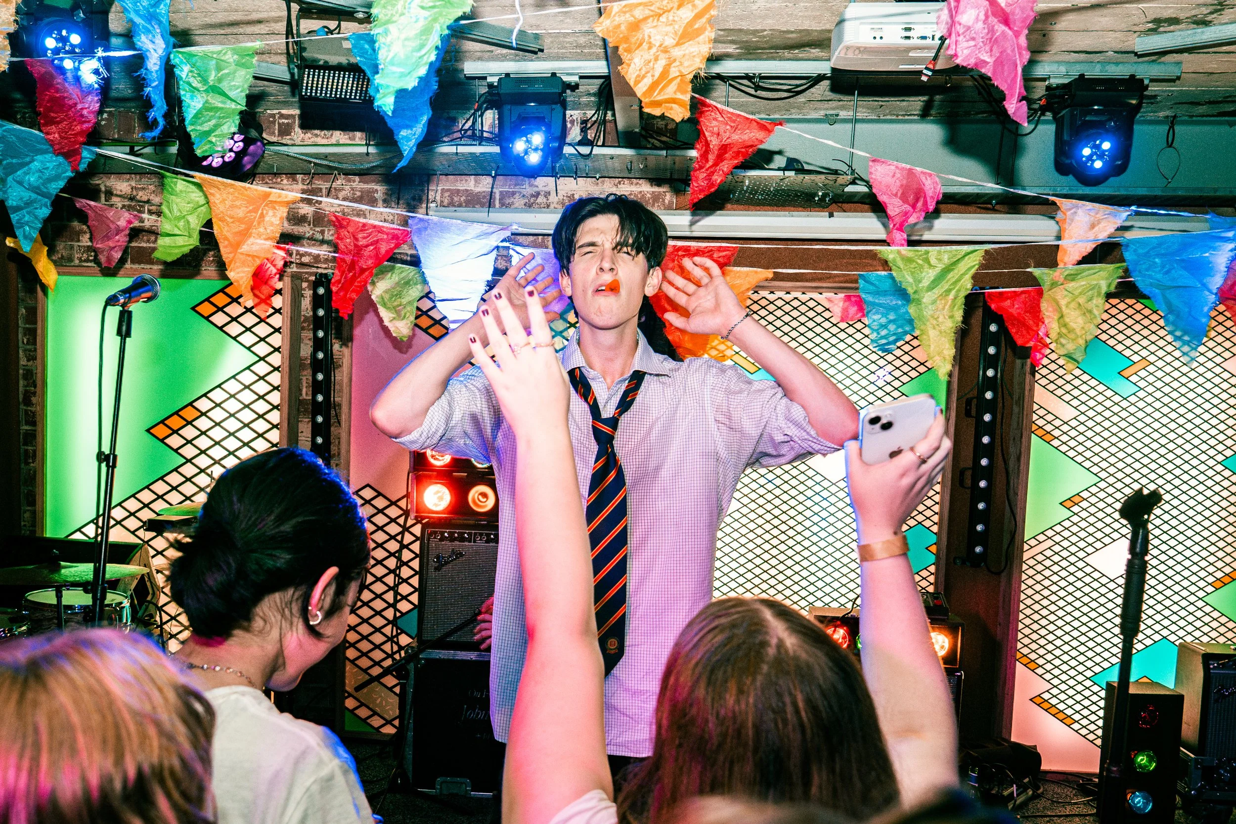 A young man in a shirt and tie stands on stage at a lively party or music event with colorful bunting and stage lights, singing and dancing with his eyes closed, while people in the audience sing along and hold up their phones.