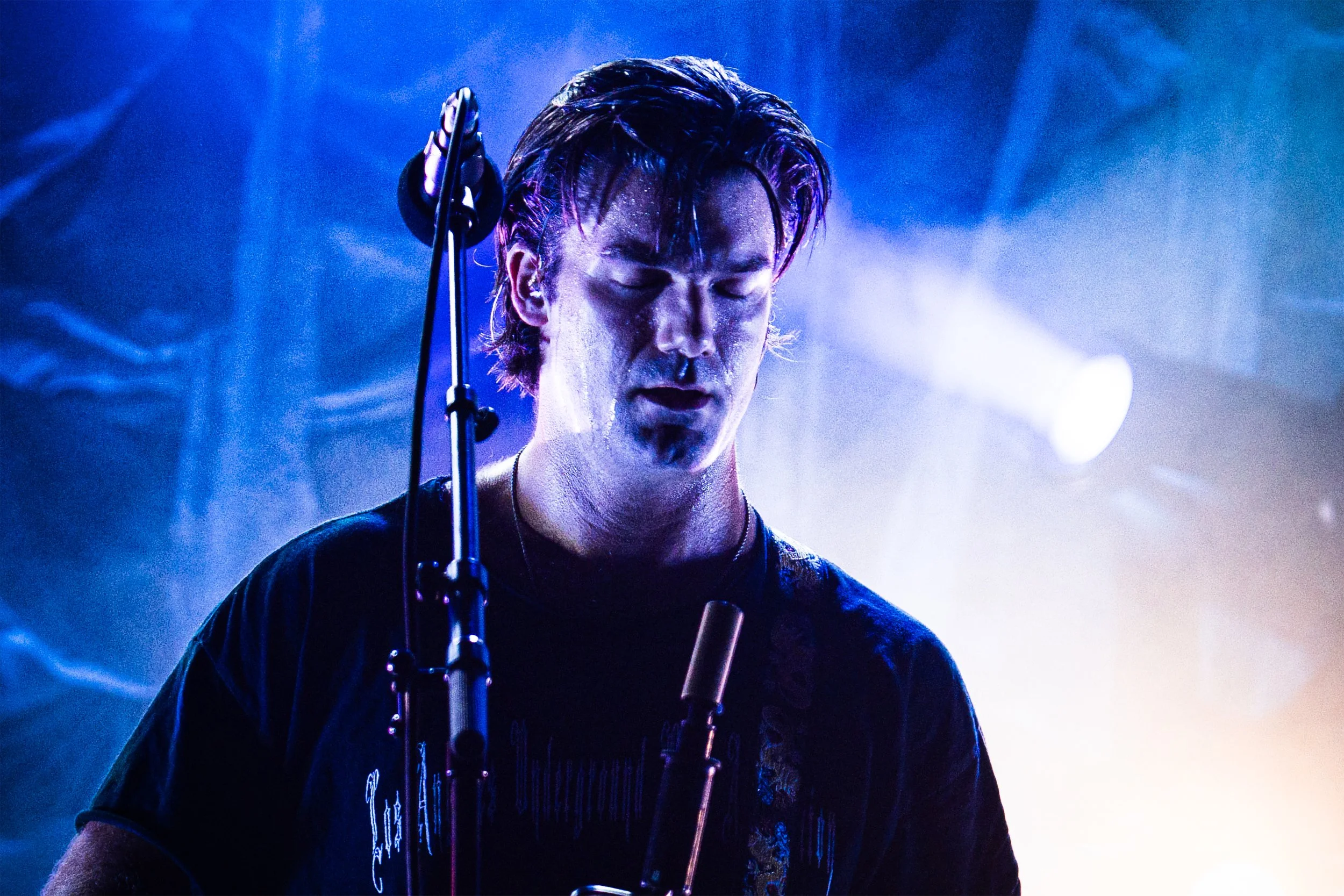A male musician on stage with eyes closed, illuminated by blue and purple stage lights, singing into a microphone.