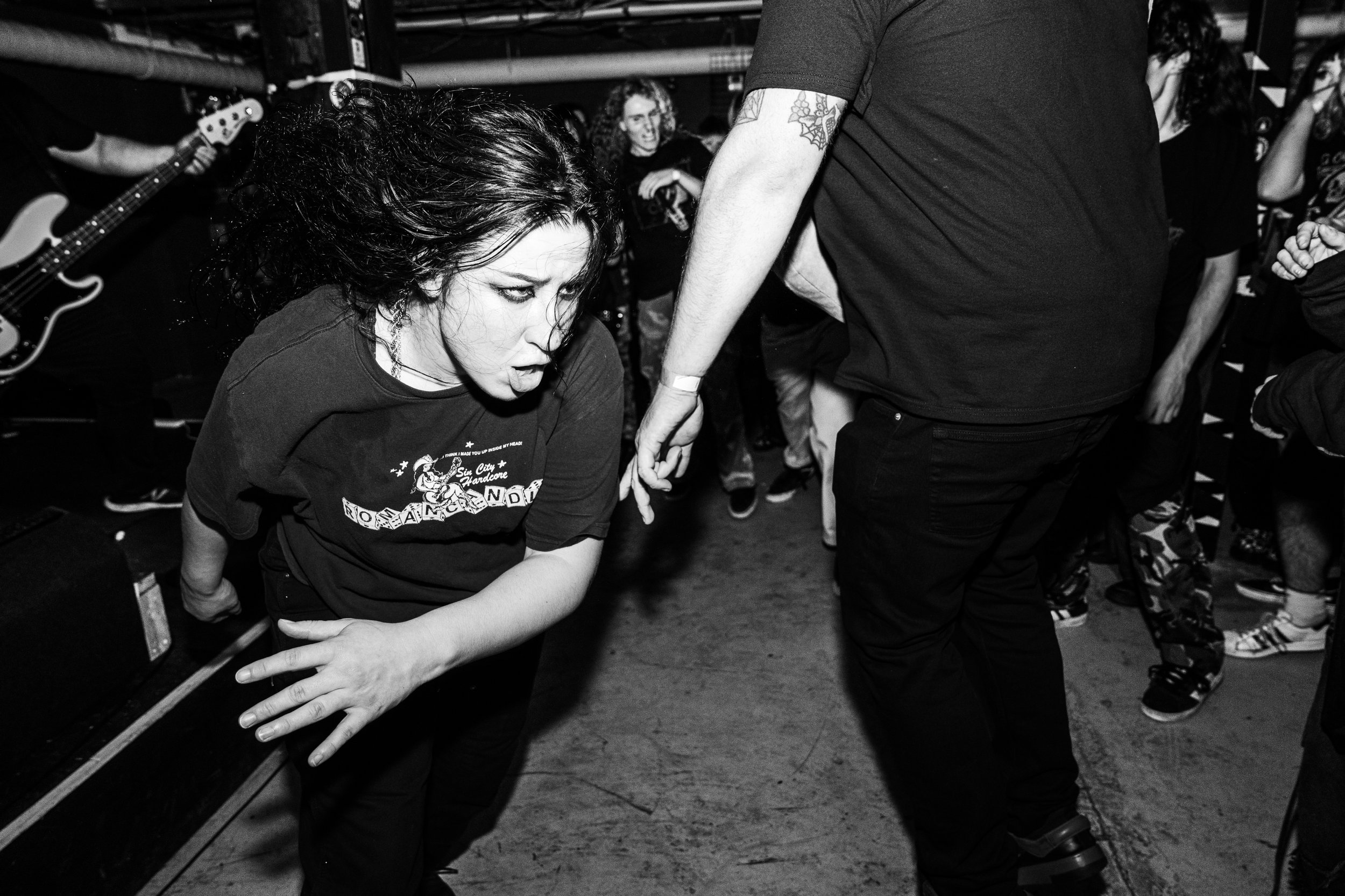 A black and white photo of a woman with dark curly hair at a live concert, leaning forward with a serious expression, holding her knee, while a man in a black t-shirt and dark pants holds her hand or wrist.