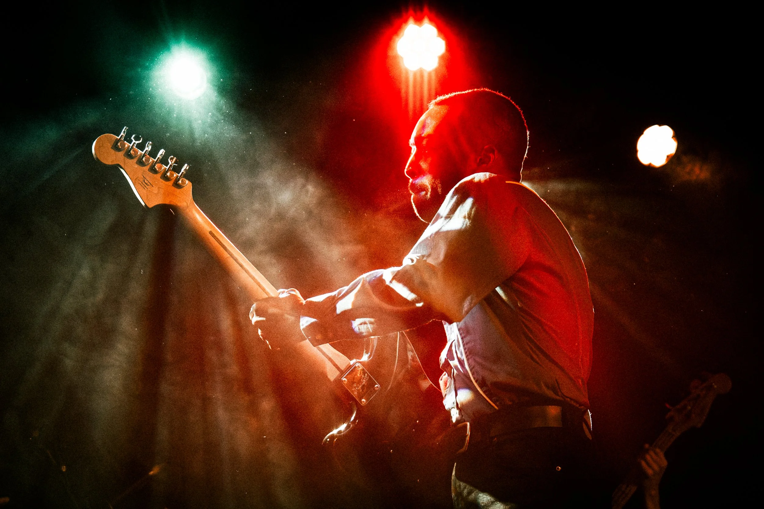 Musician playing electric guitar on stage under red and green stage lights, partially illuminated by spotlight.