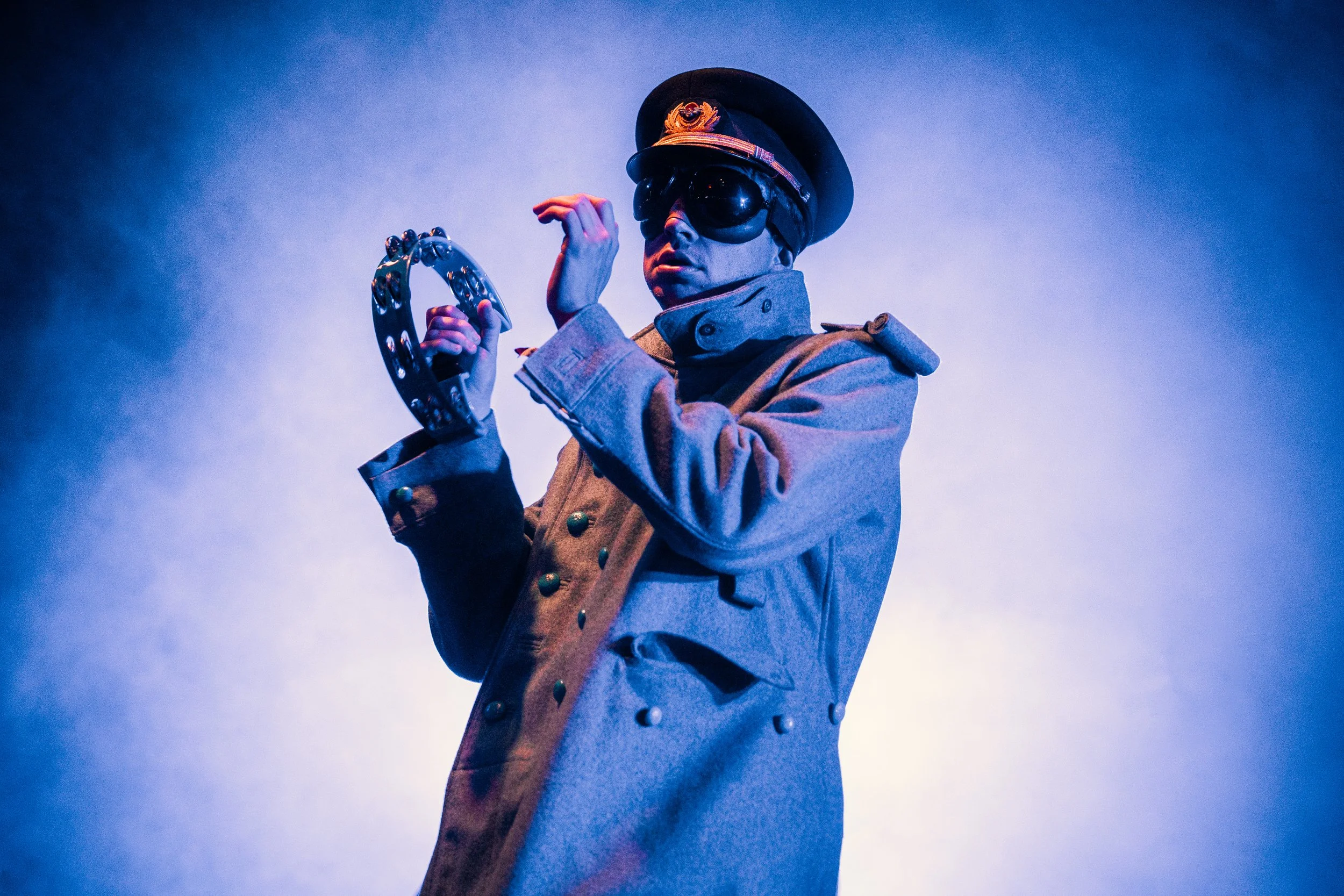 Person dressed as a military officer wearing a hat, sunglasses, and a coat, holding a horseshoe-shaped object, with dramatic lighting and a blue background.