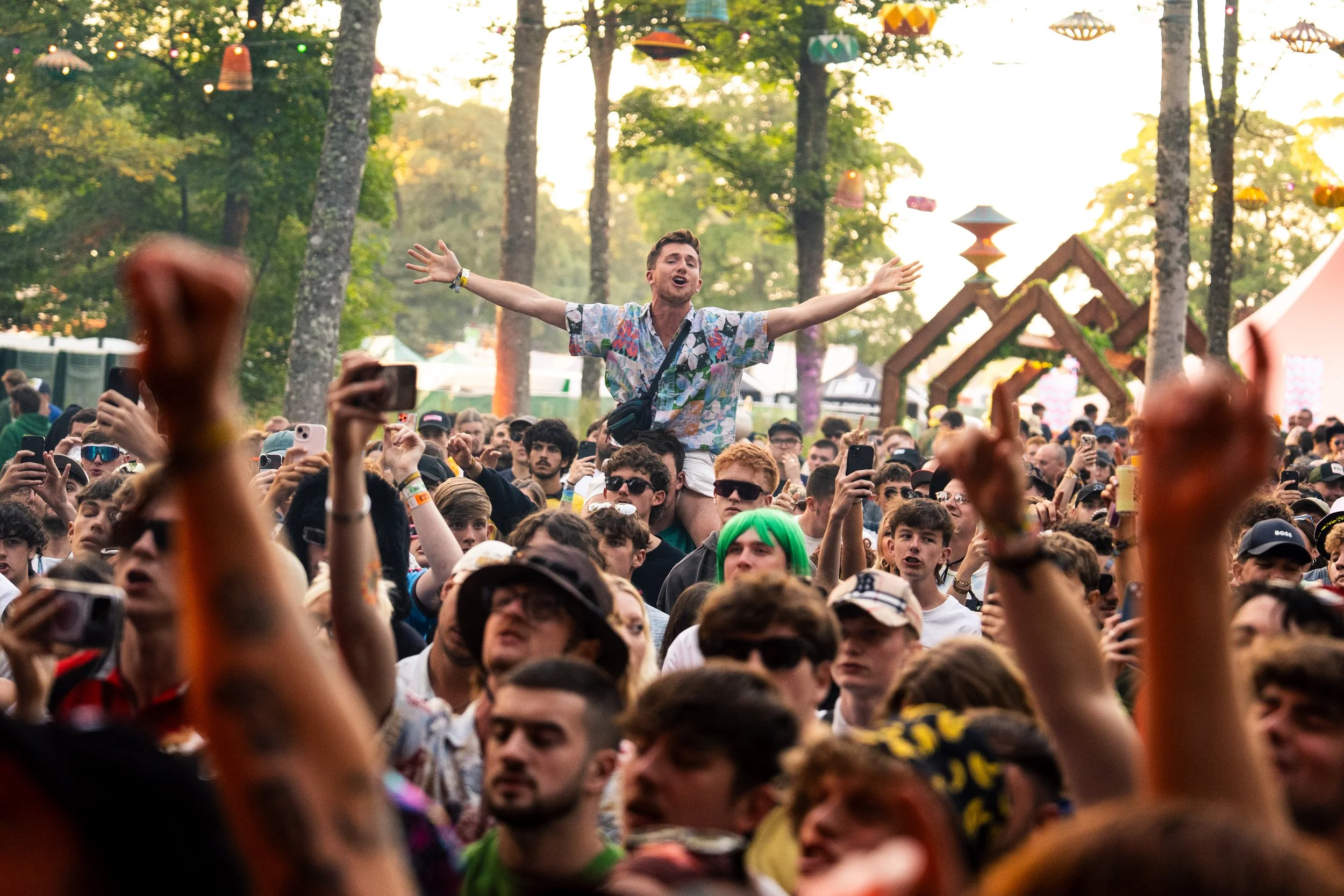 A crowd of people at an outdoor music festival, with a man sitting on someone's shoulders in the center, arms outstretched. Some attendees are holding phones, and the background features trees, decorative hanging lanterns, and festival structures.