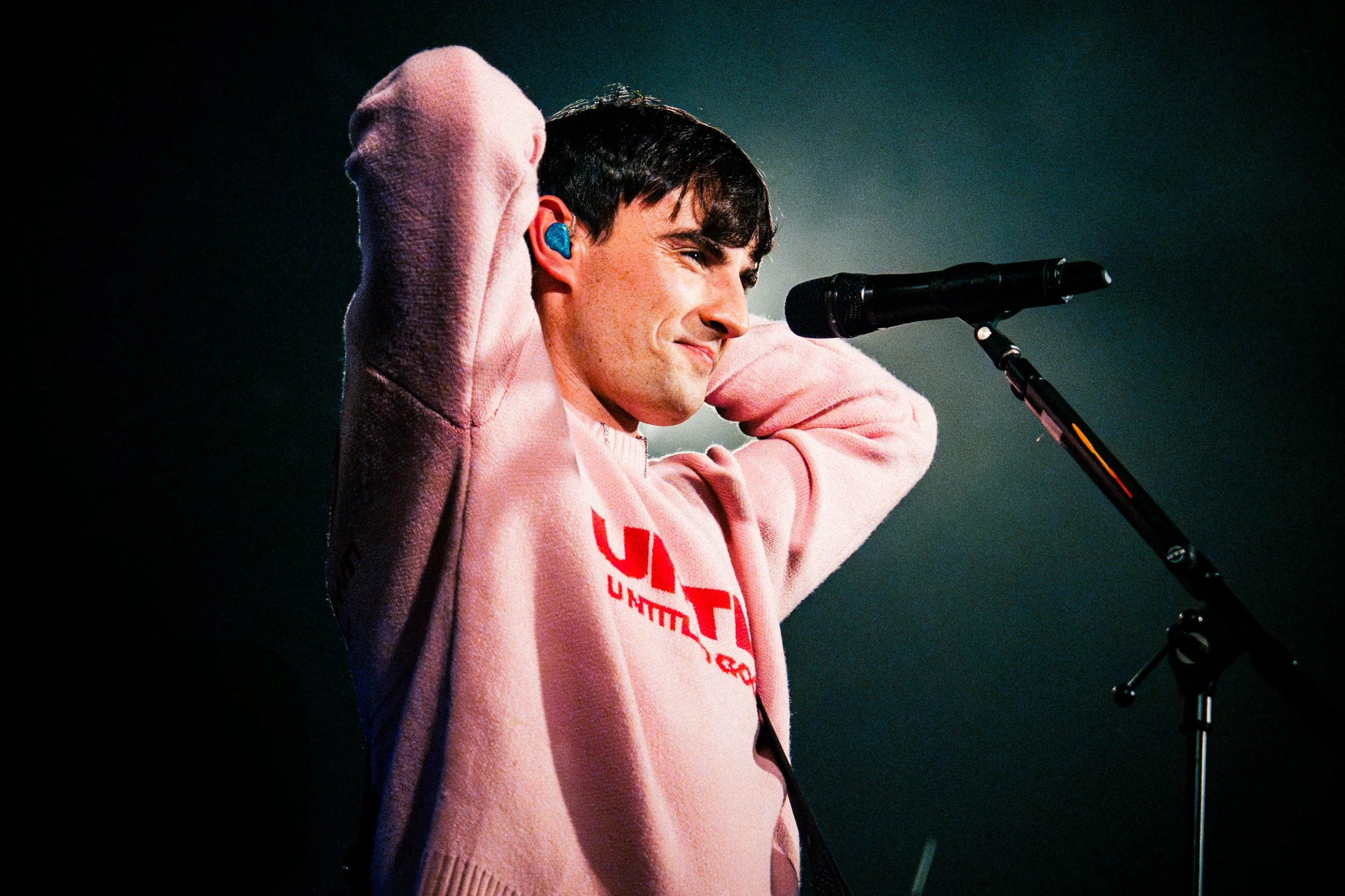 A young man performing on stage, adjusting his hair, standing in front of a microphone, wearing a pink hoodie with text on it.