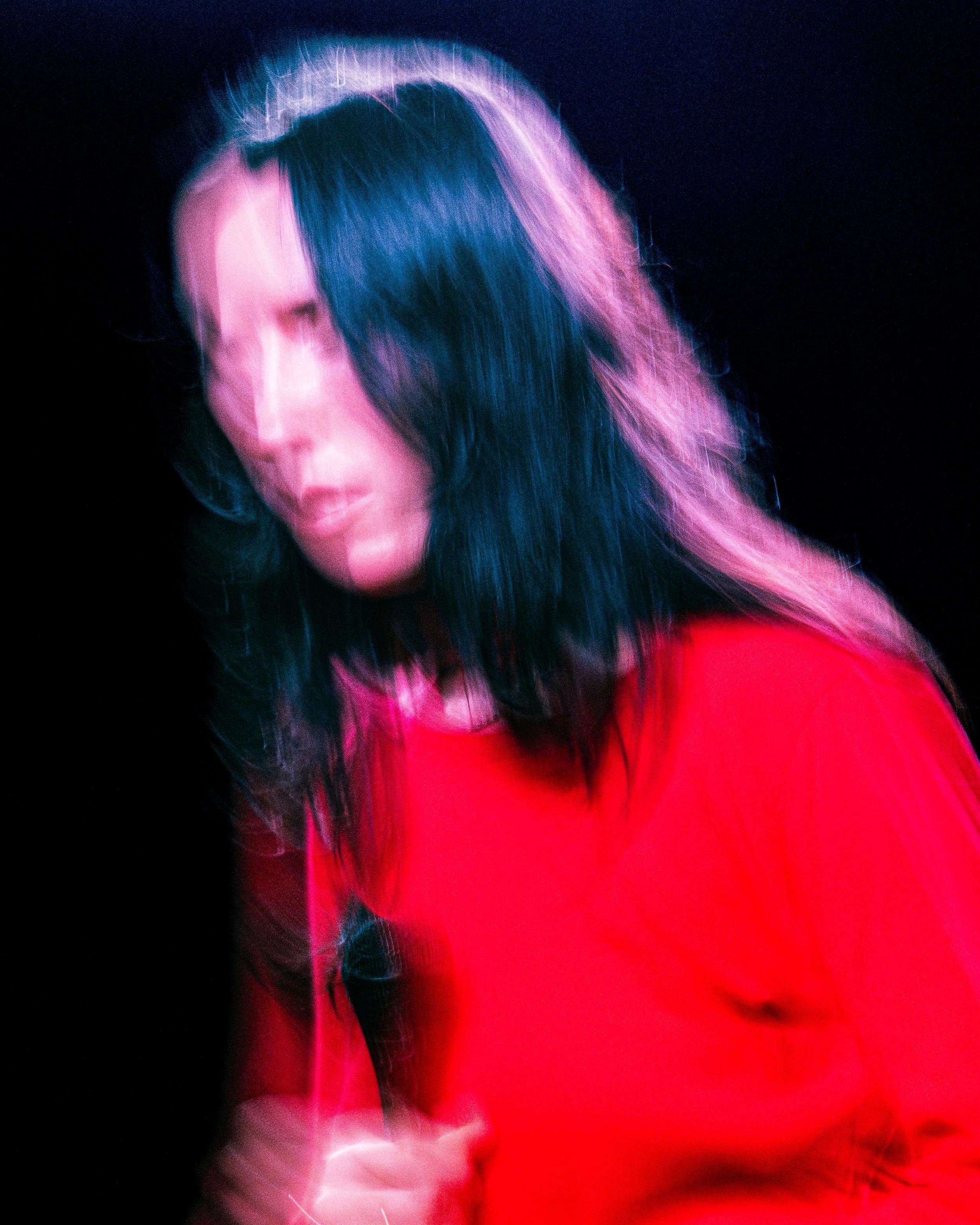 Blurry photo of a woman with dark hair wearing a red shirt, holding a microphone, with motion blur and colorful lighting.