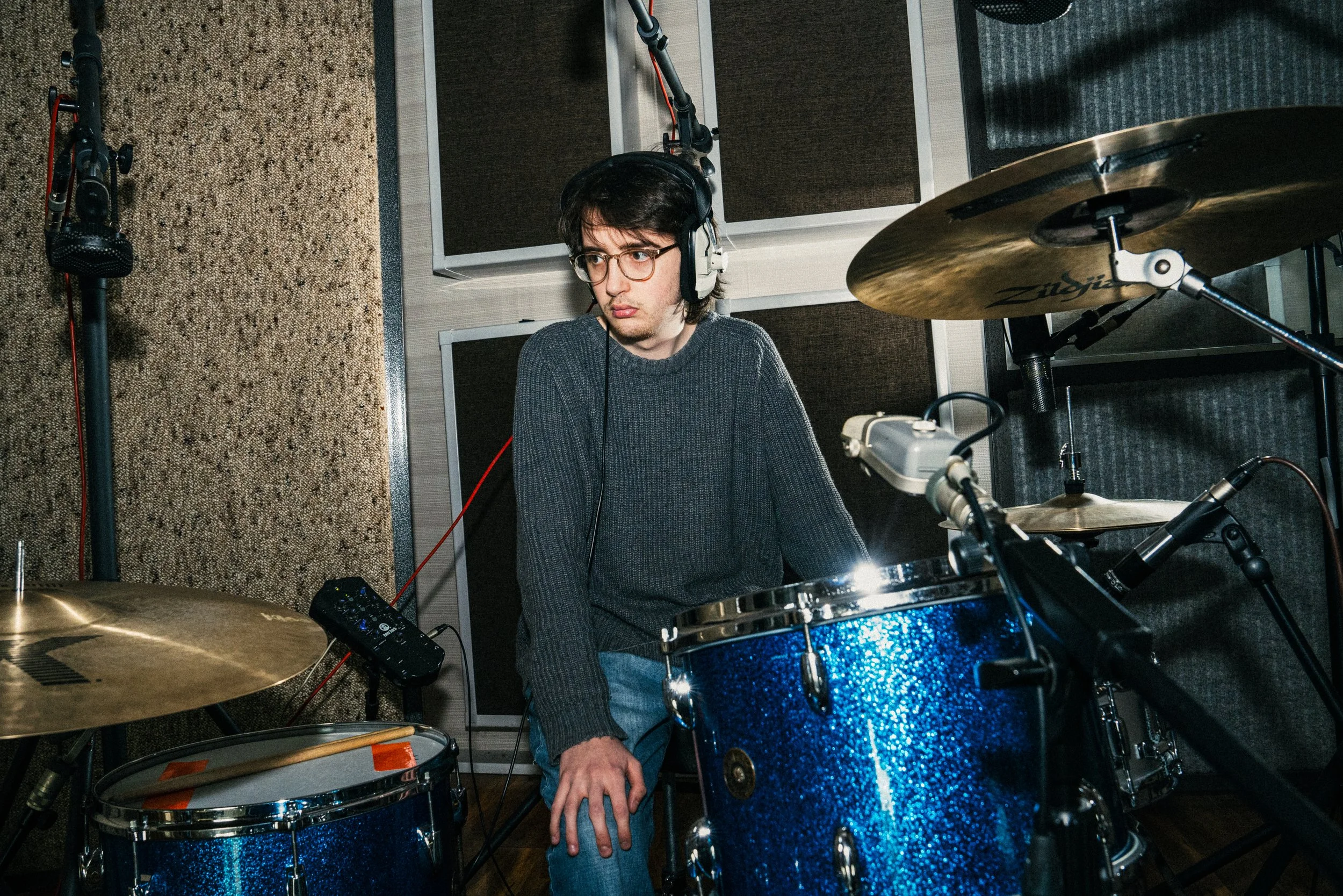 Young man with glasses and headphones sitting behind a blue drum set in a music studio.