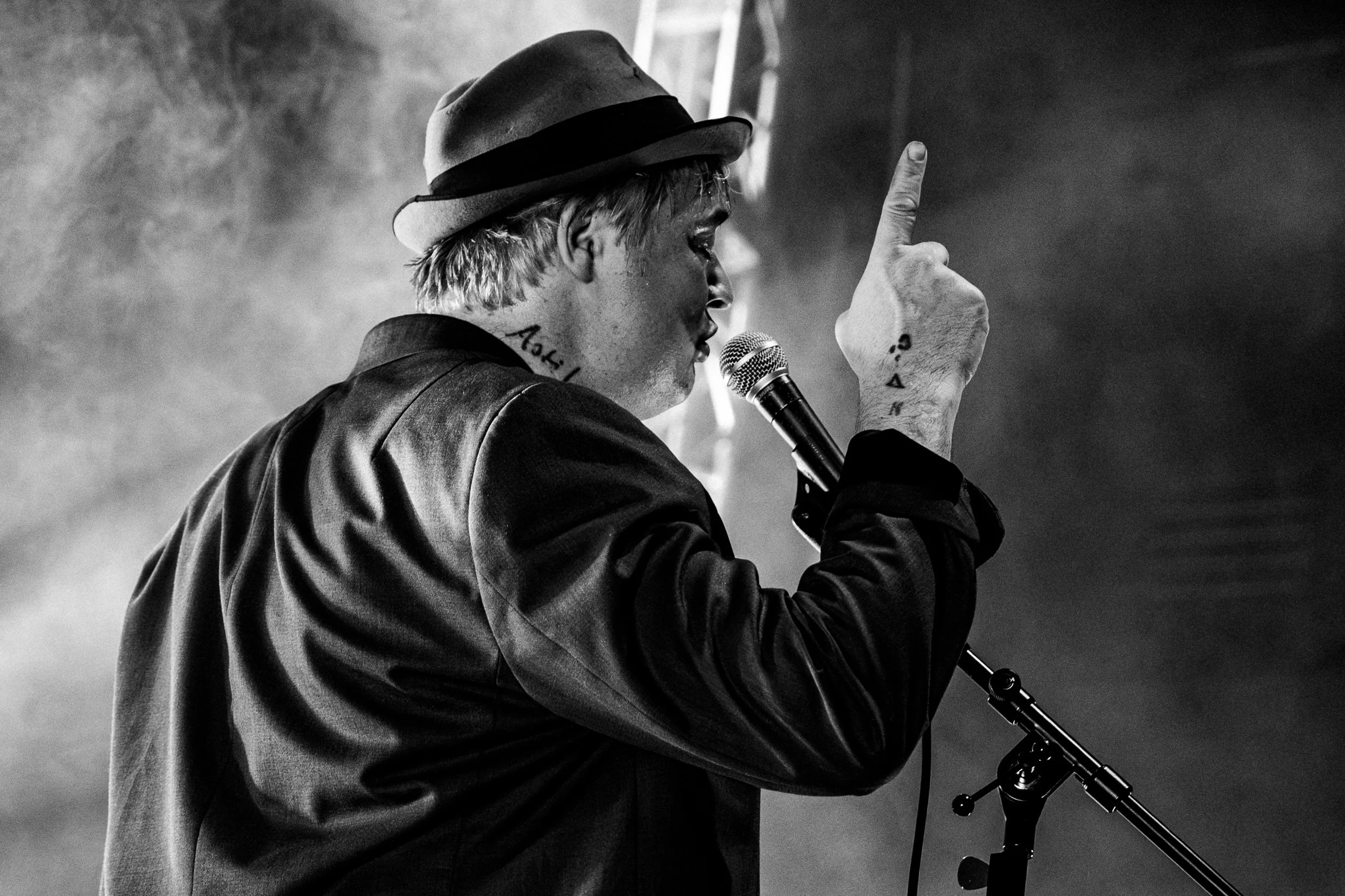 A man with tattoos on his neck and hand wearing a fedora hat and black leather jacket, singing into a microphone, with one finger raised.