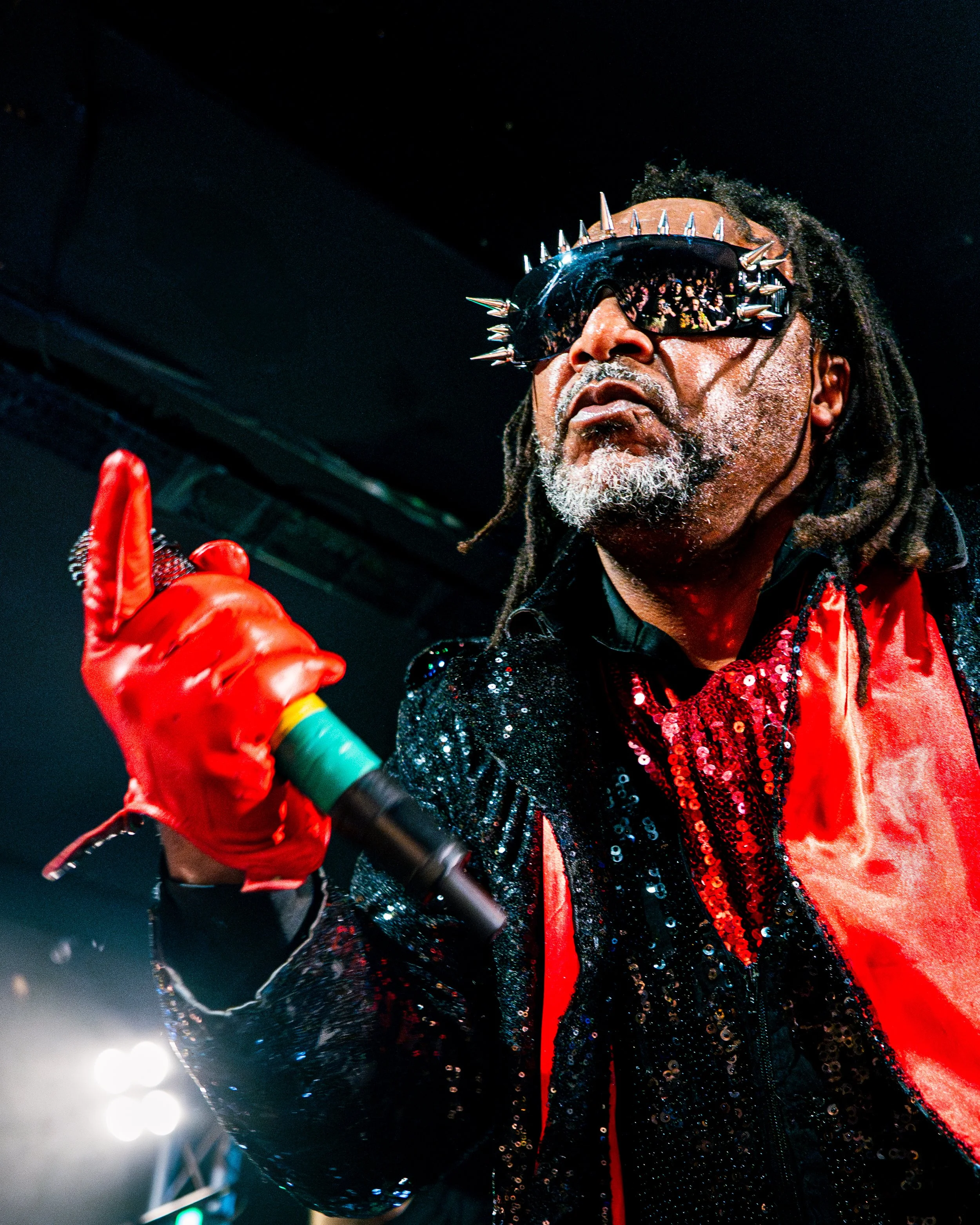A performer with dreadlocks wearing spiked sunglasses, a red and black sequined jacket, and red gloves, holding a microphone during a concert.