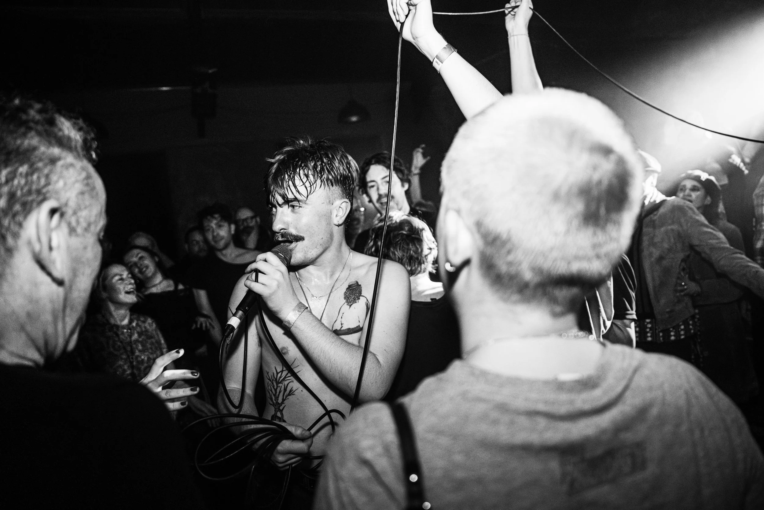 A black and white photo of a shirtless man with a mustache singing into a microphone at a party or concert, surrounded by people dancing and enjoying the music. GANS