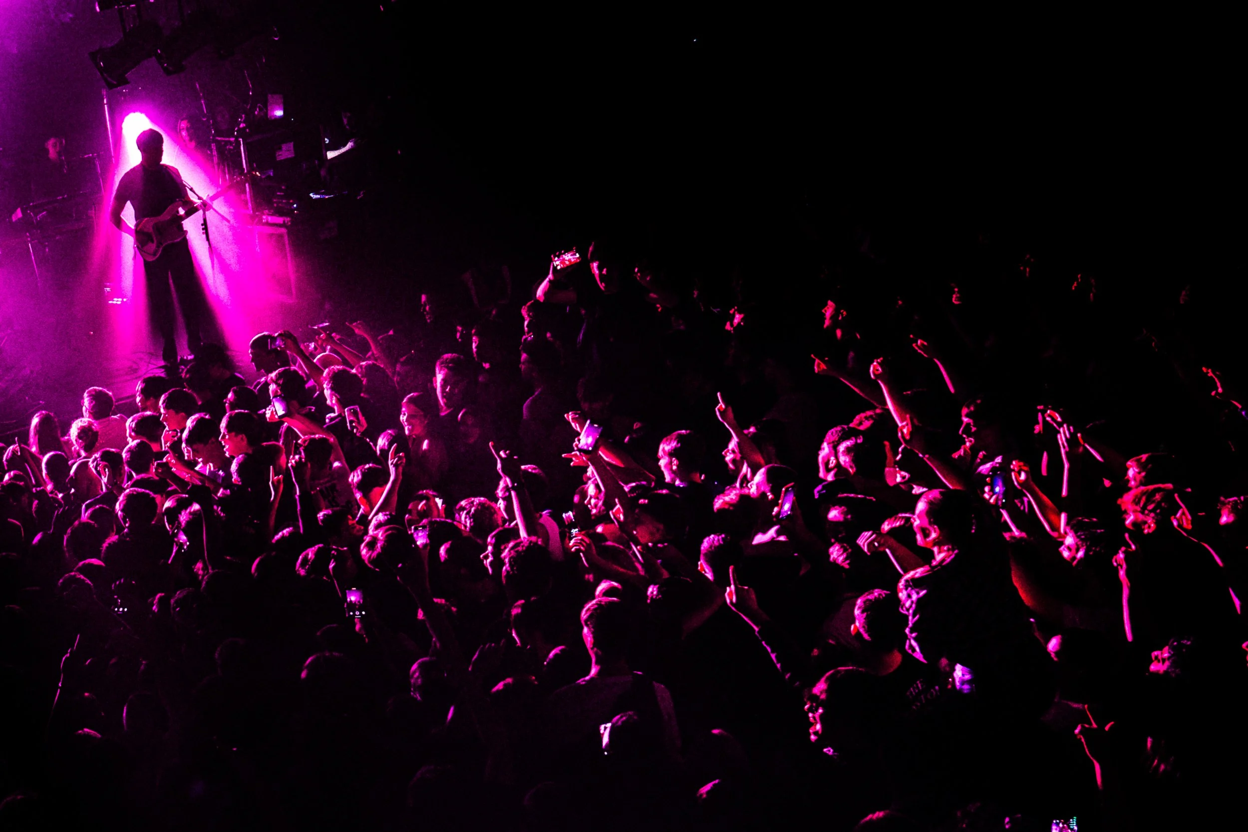 Crowd of people at a concert, with many holding up their phones, illuminated by purple stage lights, and a musician playing guitar on stage