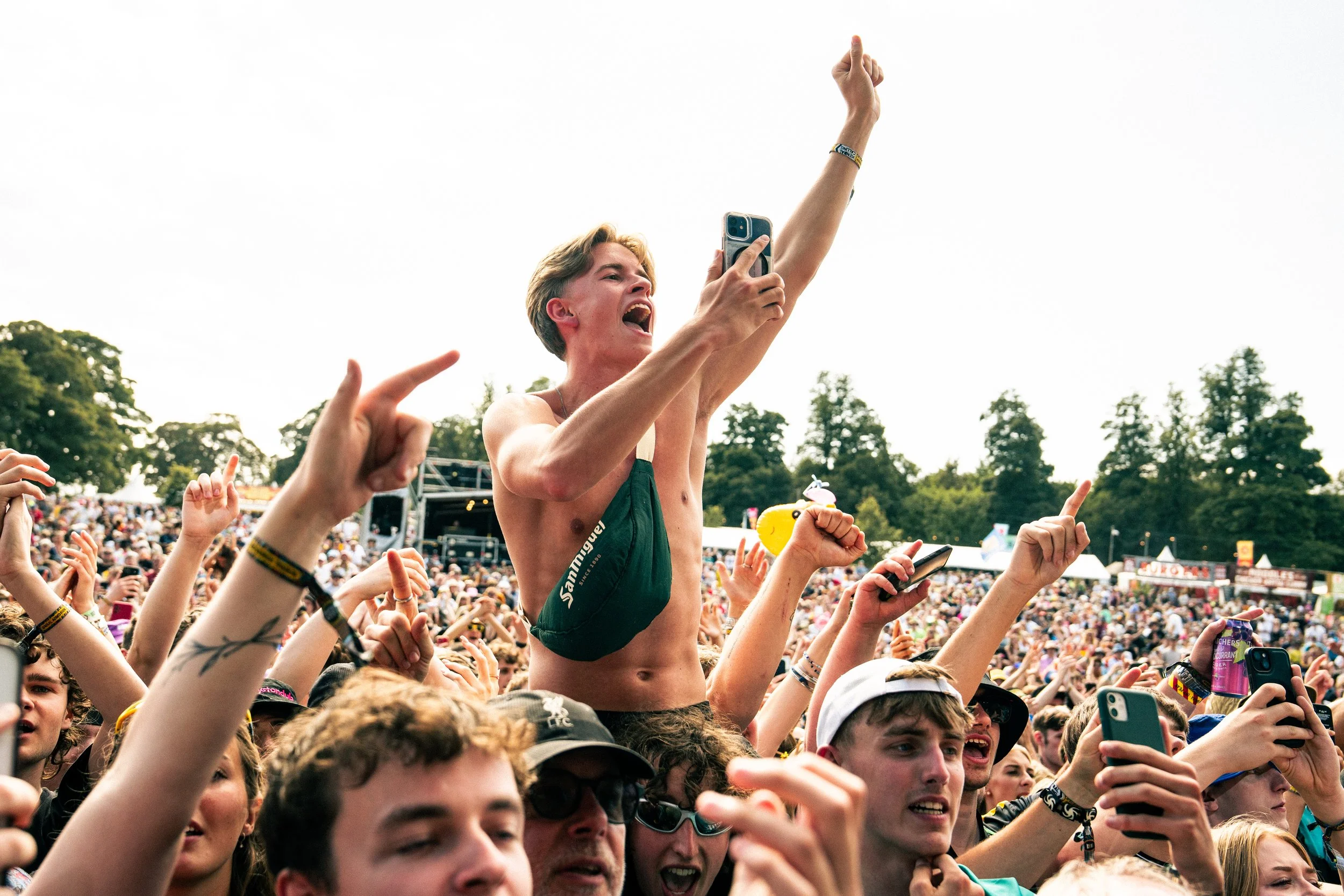 A young man with short blonde hair and a green swimsuit is crowd surfing at an outdoor concert, holding a smartphone in one hand and raising his other arm. The audience around him is cheering and taking photos.