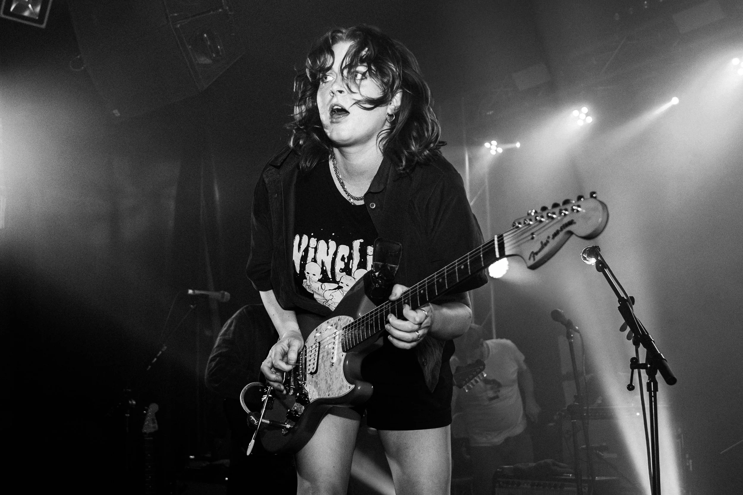 A female guitarist on stage during a live music performance, with her hair partially covering her face, wearing a dark t-shirt and shorts, holding an electric guitar, and surrounded by stage lighting and microphones.