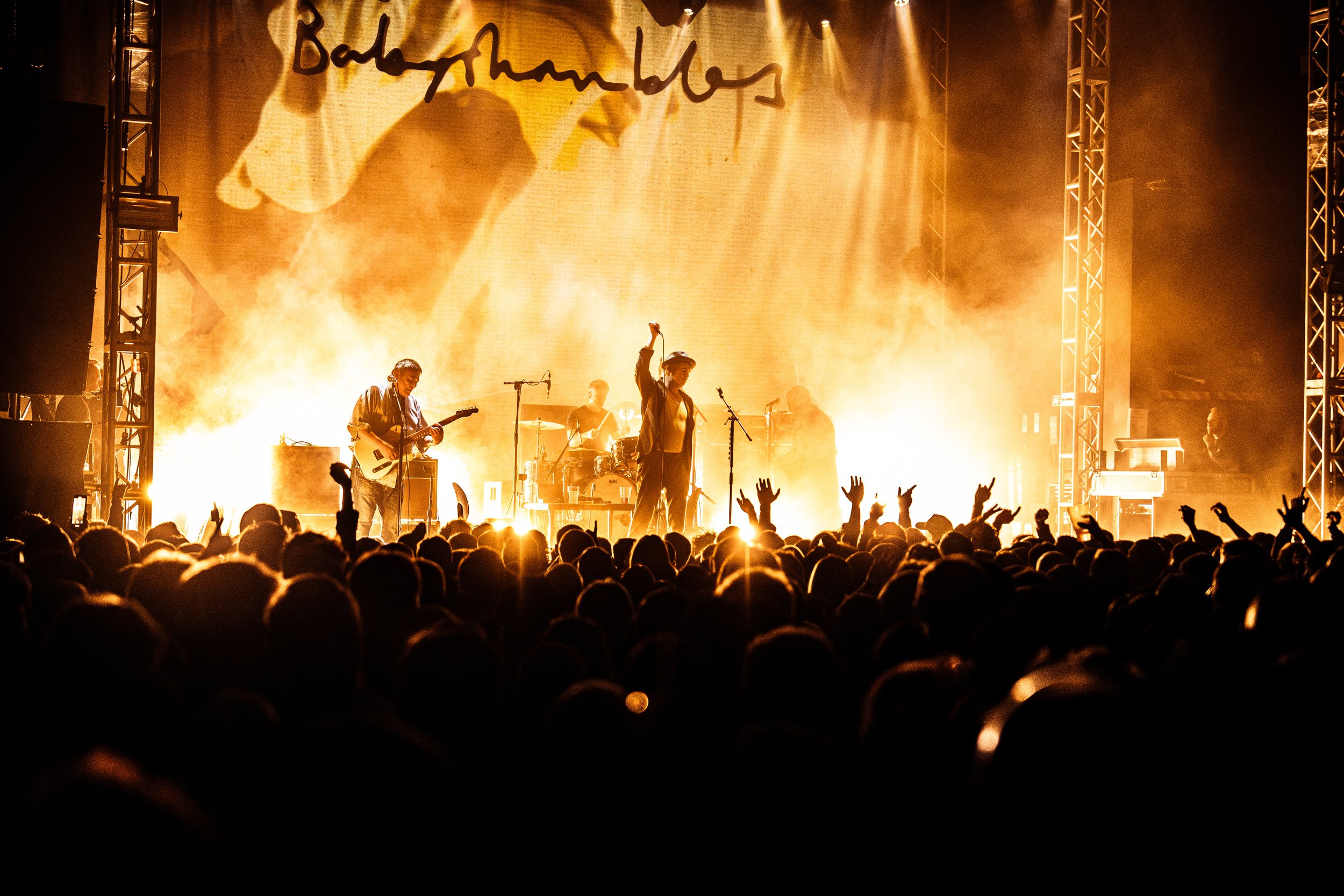 A live band performing on stage with bright yellow and orange lighting, silhouettes of the audience with hands raised, and a large backdrop with partially visible text.
