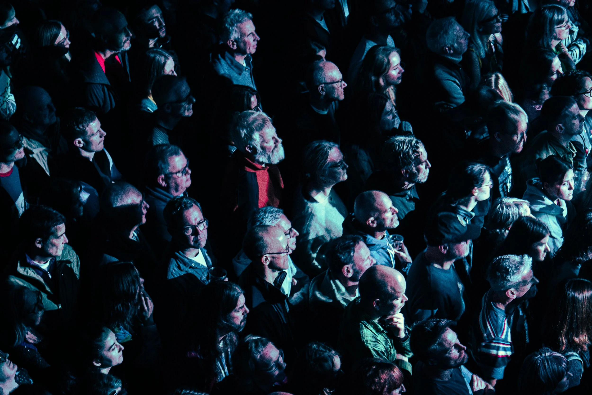 A crowd of people standing closely together at a concert or event, illuminated by blue stage lighting, facing towards the stage.