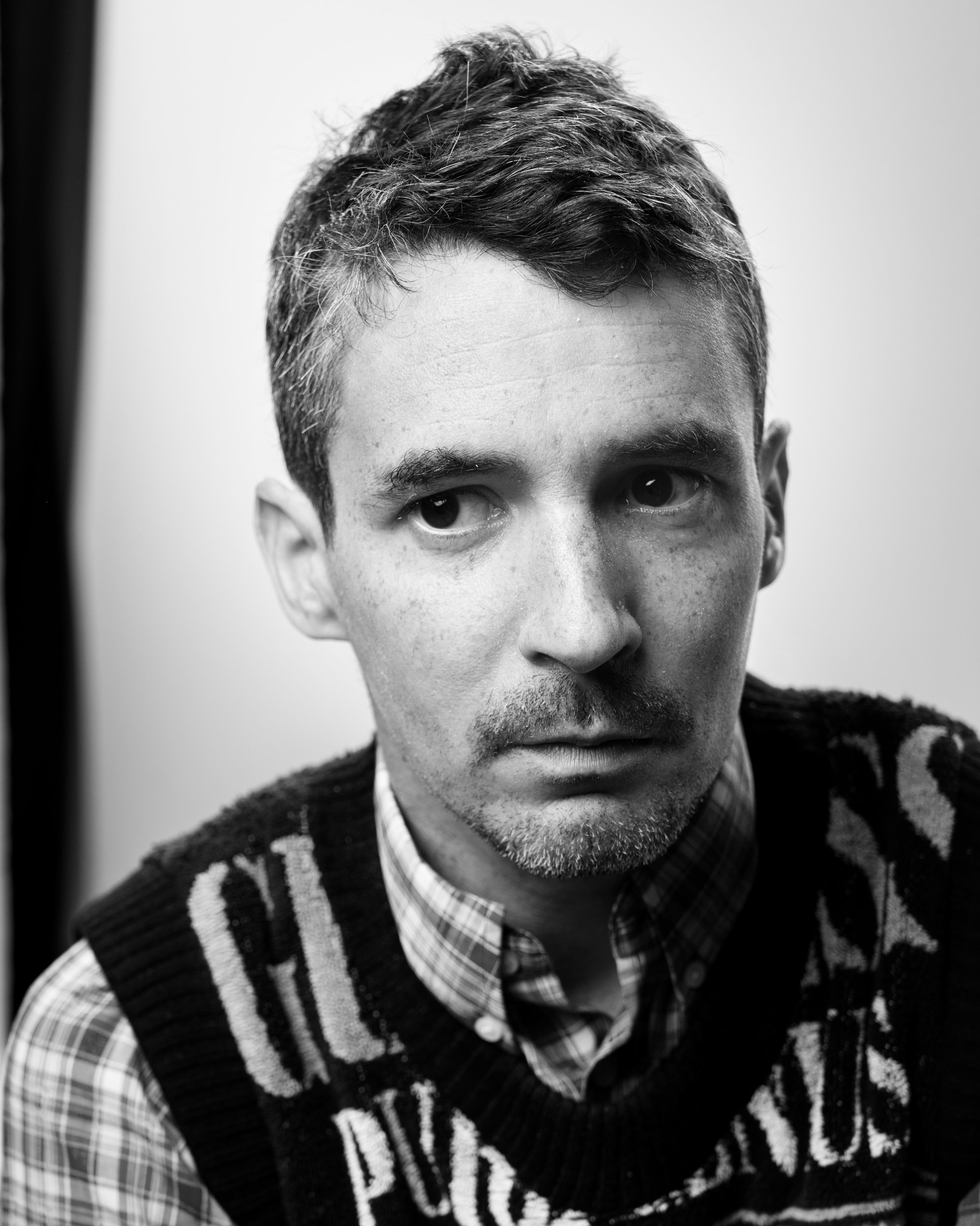 Black and white close-up portrait of a man with short hair, slight stubble, wearing a plaid shirt and a patterned knitted vest.