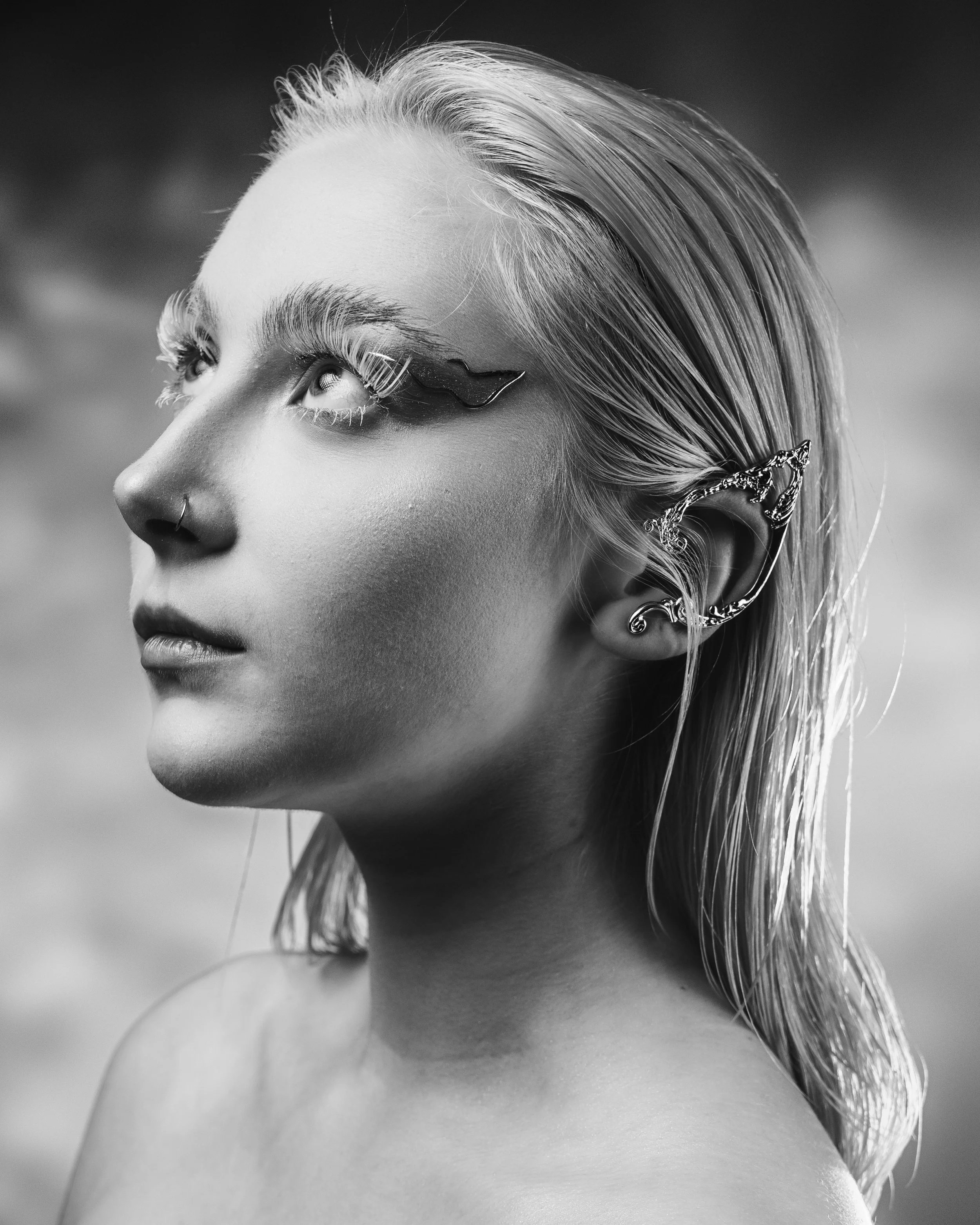 Black and white portrait of a woman with wet blonde hair, wearing artistic eye makeup and multiple earrings, looking upwards.